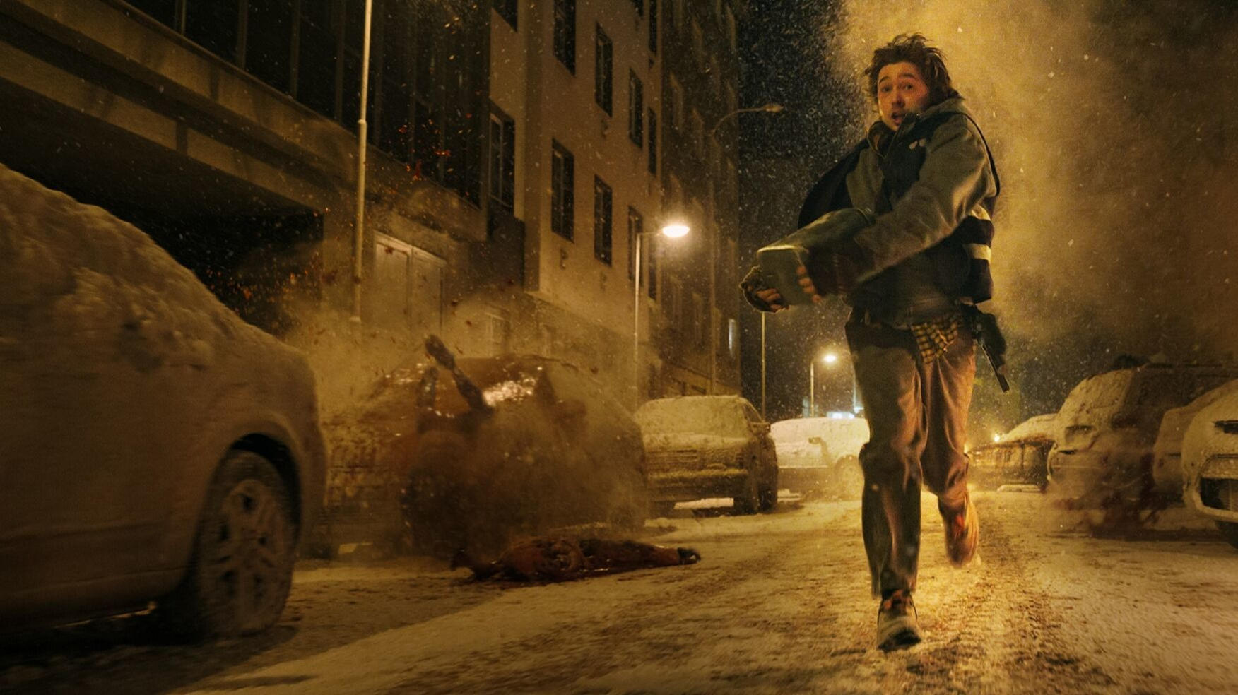 Ein Mann rennt im Schnee durch eine Straße, während im Hintergrund Autos in Flammen stehen.