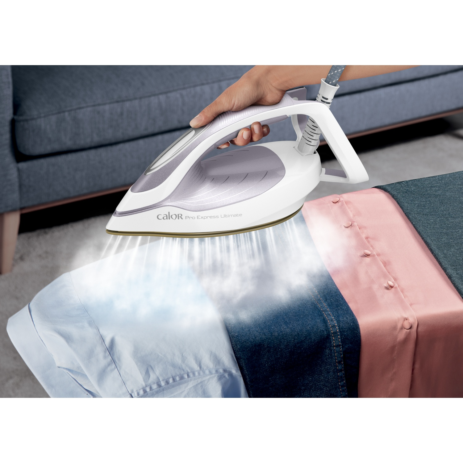 A hand holds a white and purple steam iron, releasing steam onto a blue shirt.