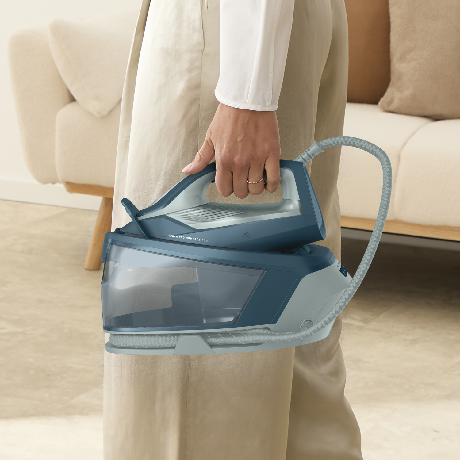 Person holding a blue steam iron station in front of a light sofa in a living room.