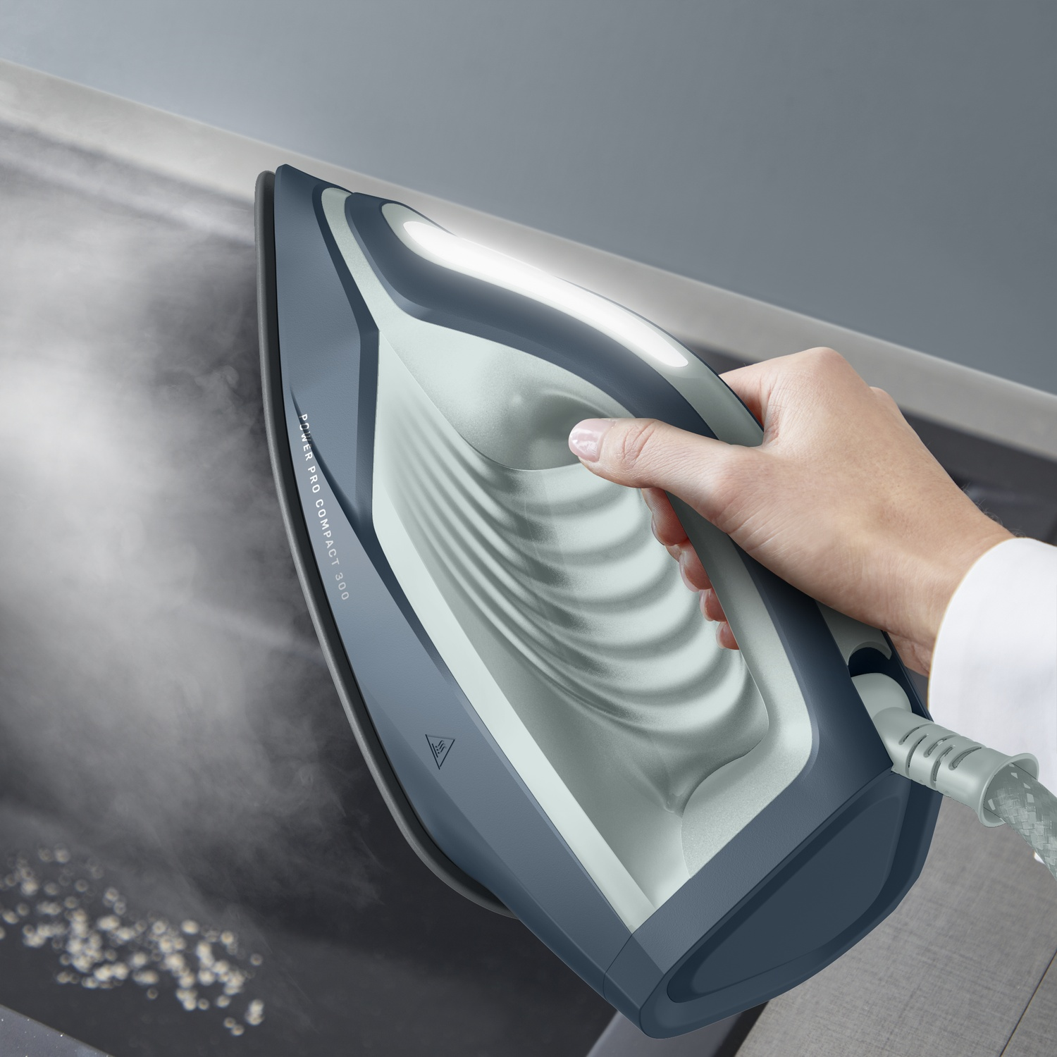 A hand holds a modern blue-gray iron, releasing steam onto a fabric surface.