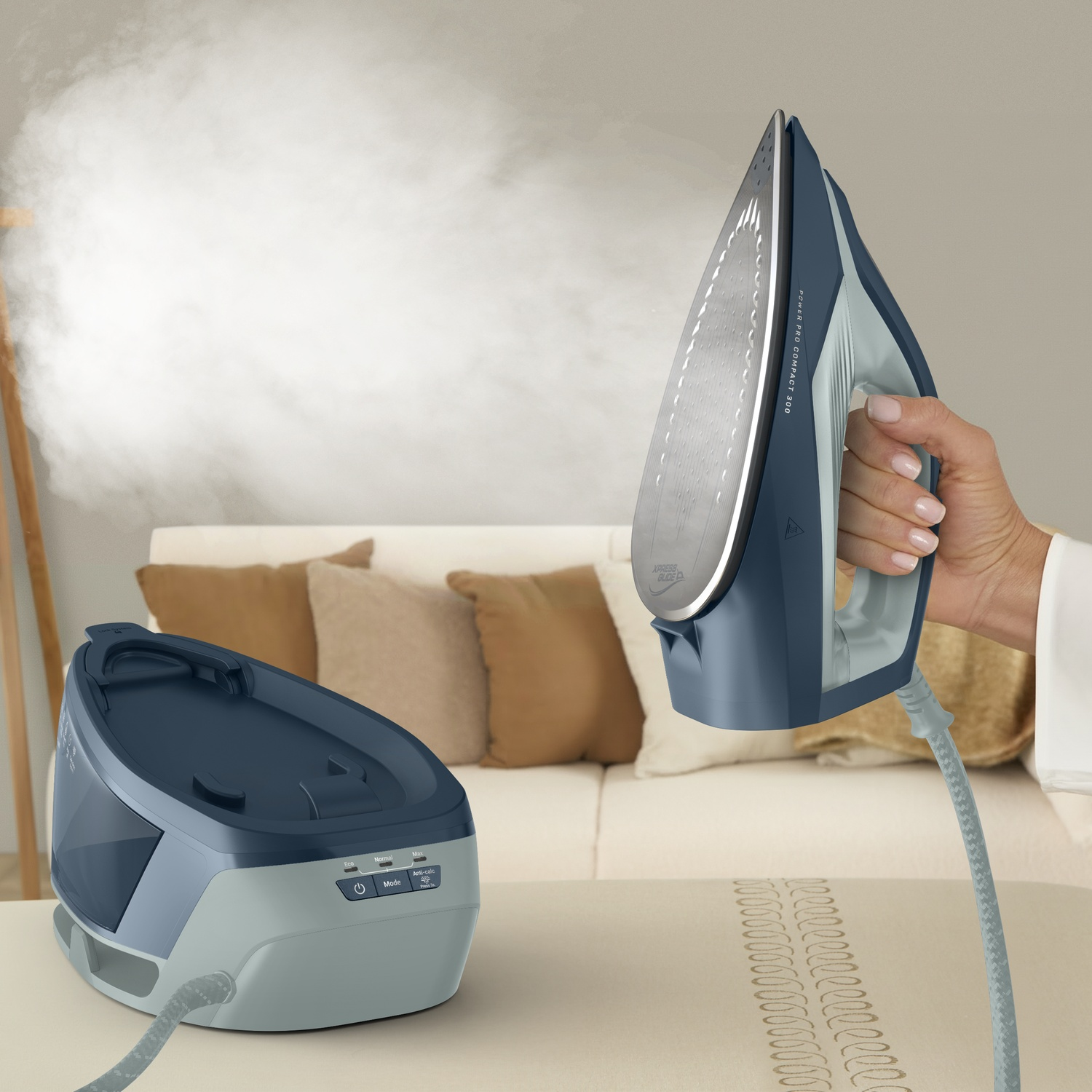 A hand holds a blue steam iron emitting a cloud of steam, with its base station on an ironing board in a living room.