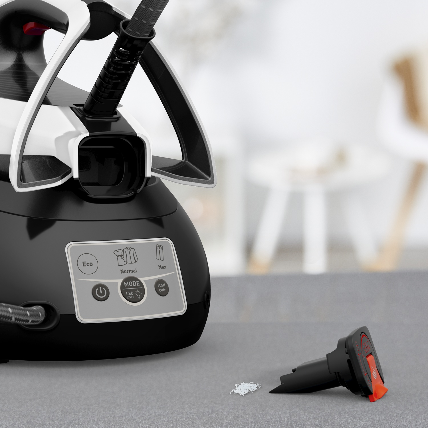 A black and white steam iron with base station stands on a table, with limescale and an attachment nearby.