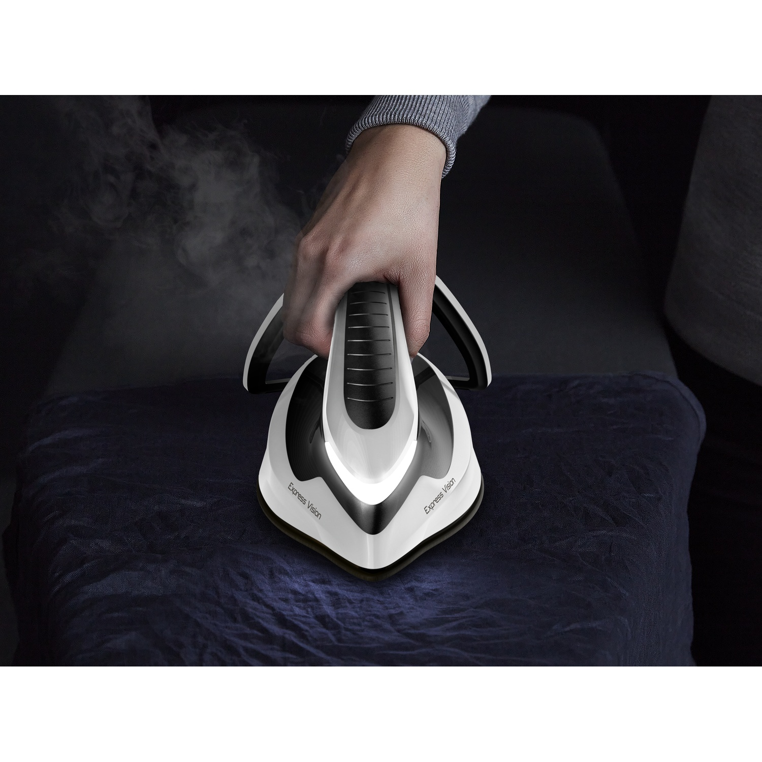 A hand holds a white steam iron releasing vapor over dark fabric.
