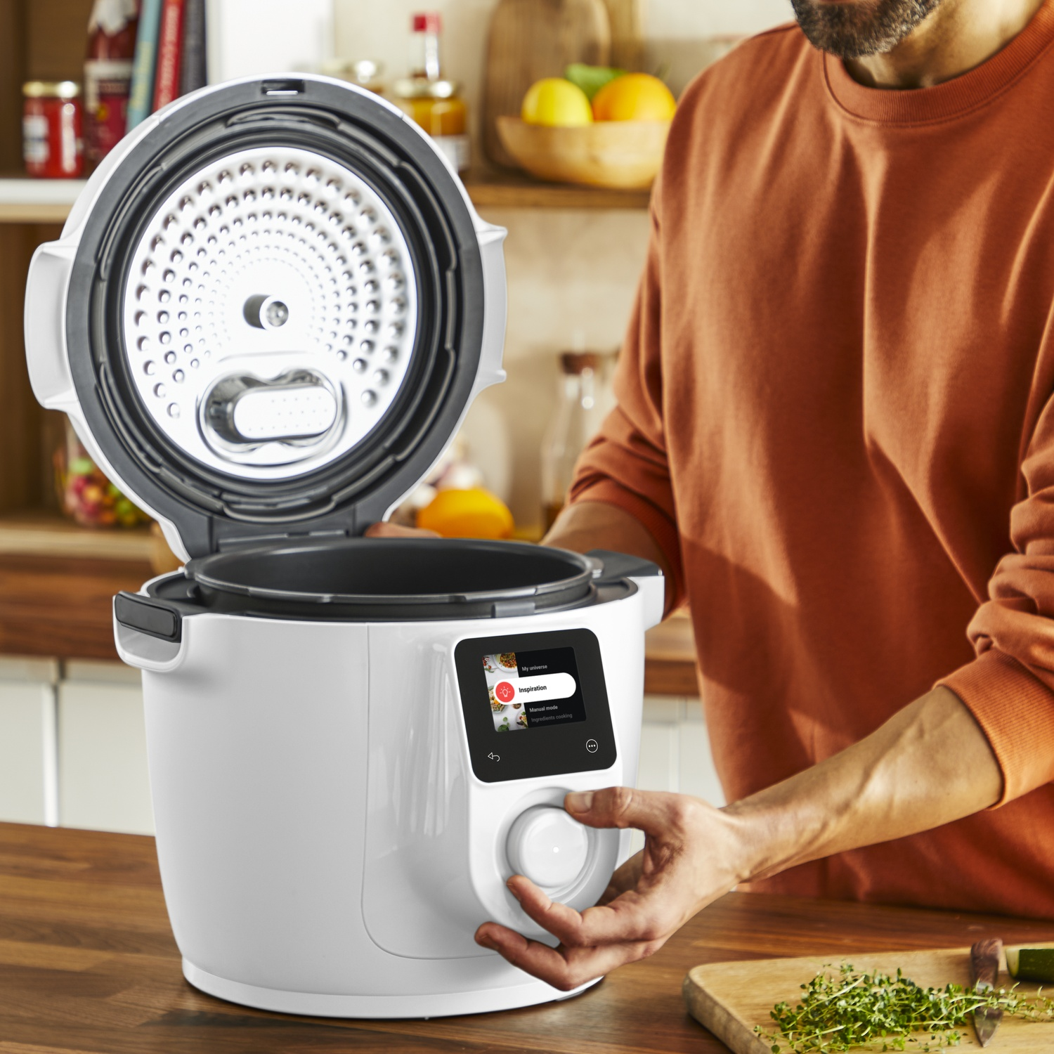 Een man in een oranje trui bedient het bedieningspaneel van een witte multicooker in een keuken.