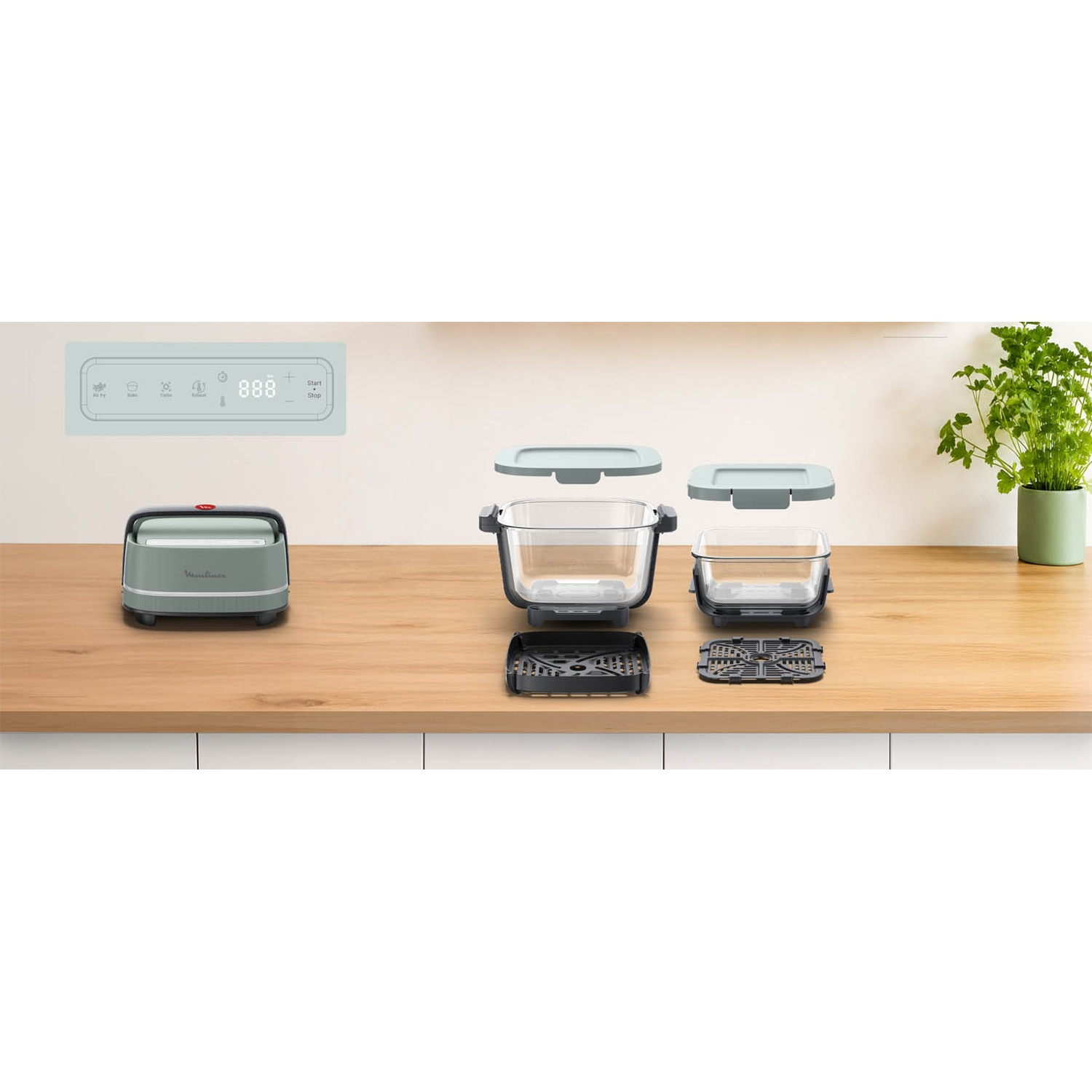 Een groene airfryer met glazen manden, accessoires en een plant op een houten aanrecht.