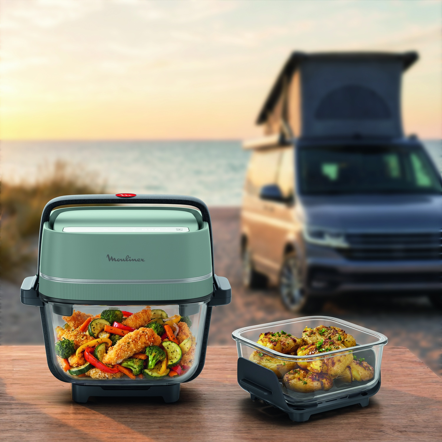 Groene Moulinex airfryer op een buitentafel, met een camper op het strand op de achtergrond.
