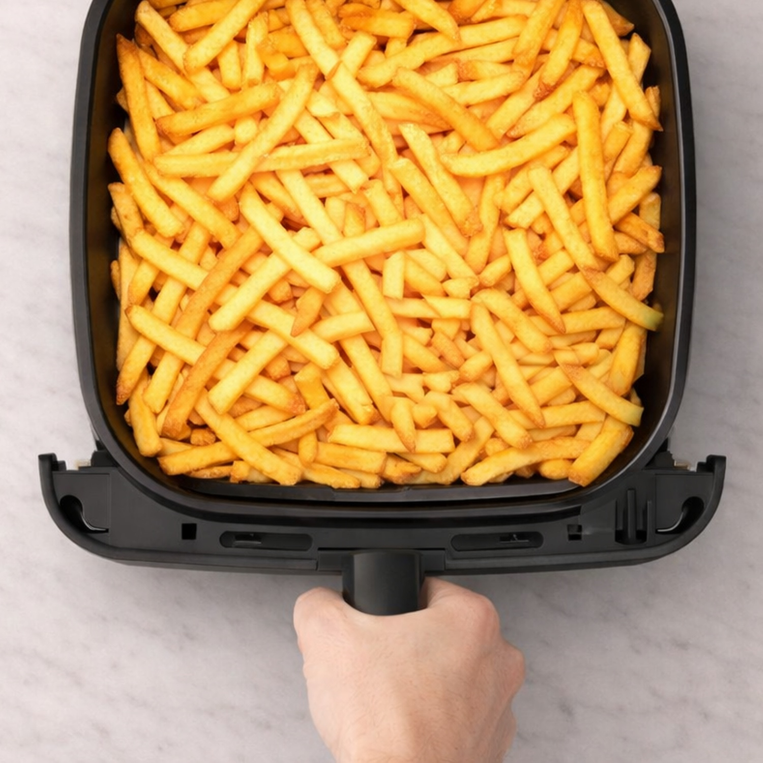 Een hand houdt de mand van een zwarte airfryer vast, tot de rand gevuld met goudbruine frietjes.