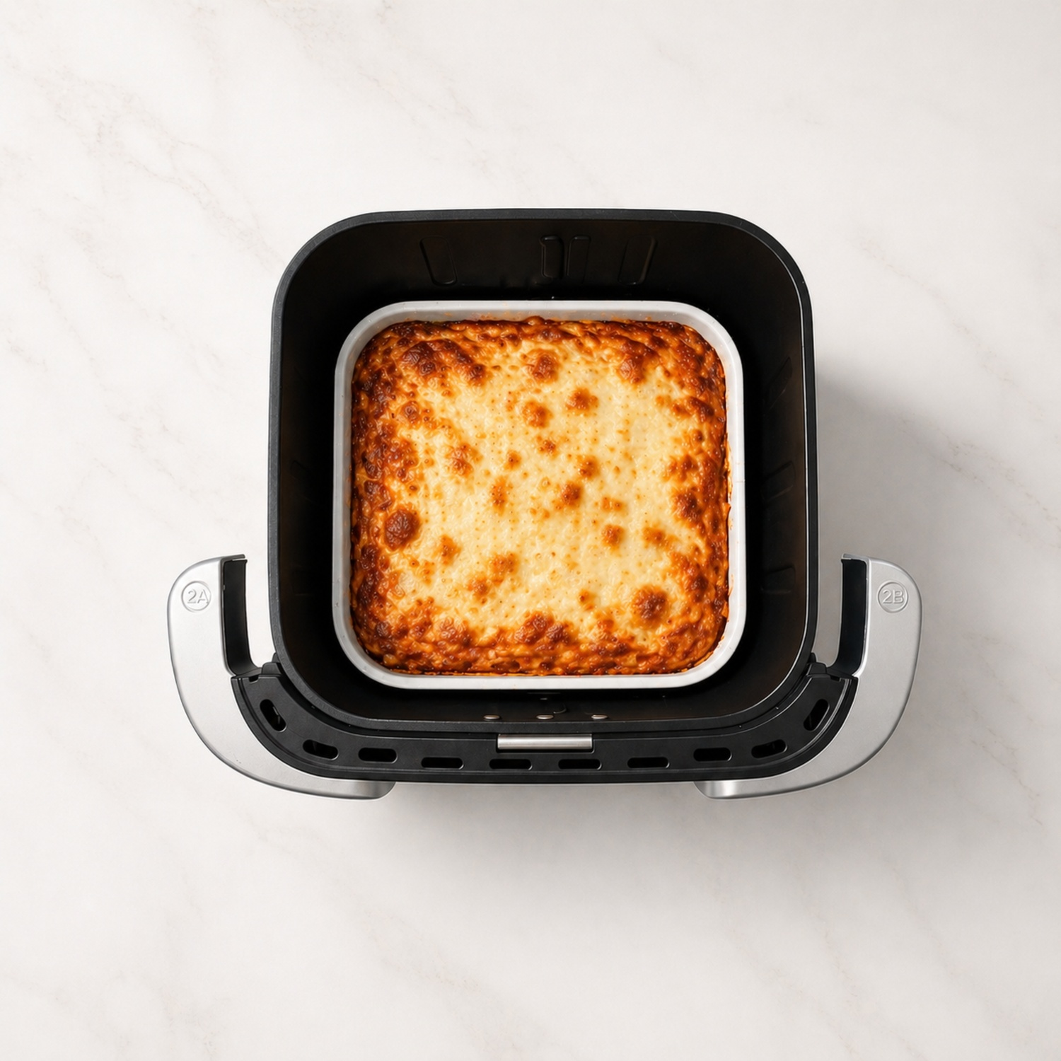 Bovenaanzicht van een gebakken gerecht in een witte ovenschaal in een airfryermand op een wit marmeren aanrecht.