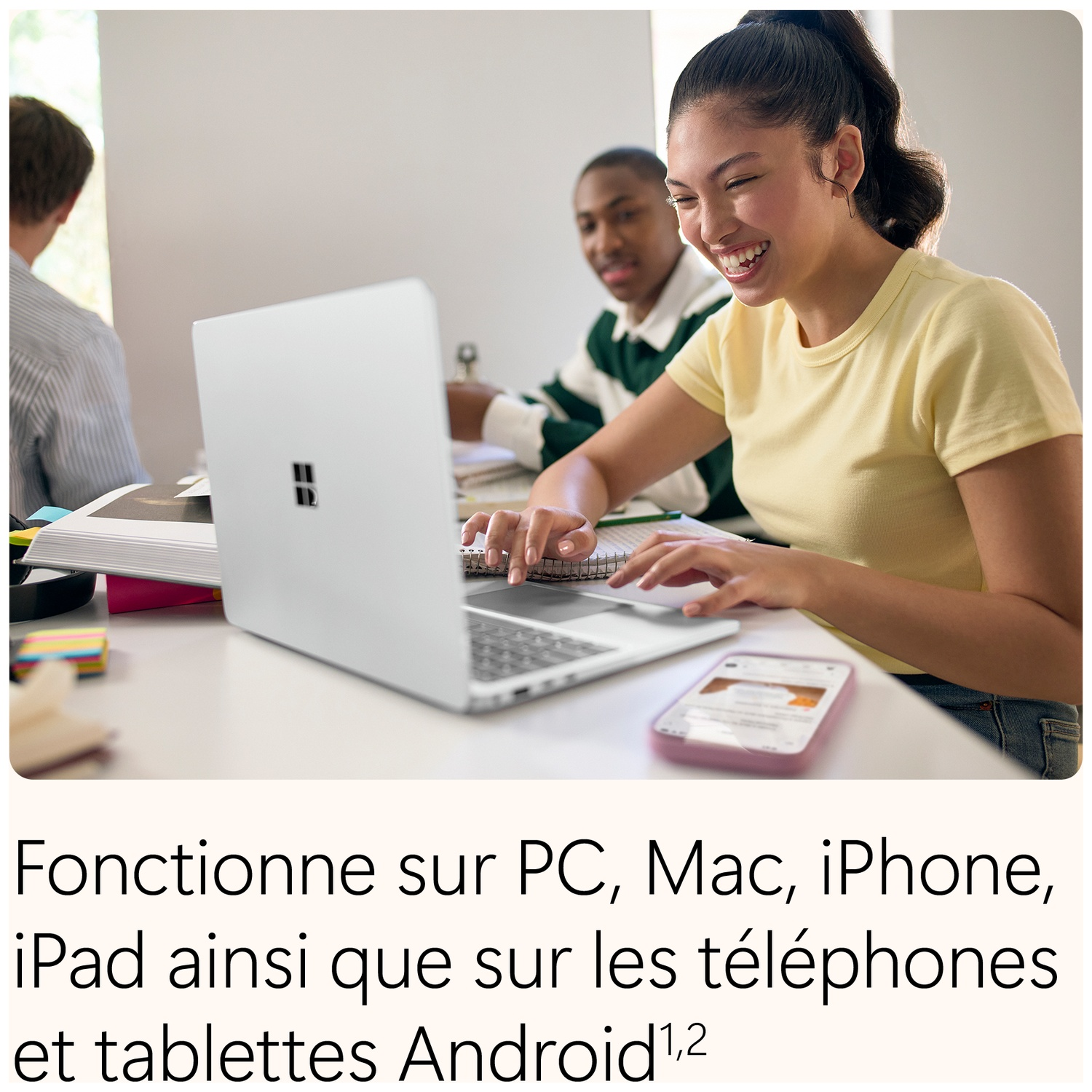 Une jeune femme souriante tape sur un ordinateur portable dans une pièce moderne, avec d'autres personnes assises à l'arrière-plan.