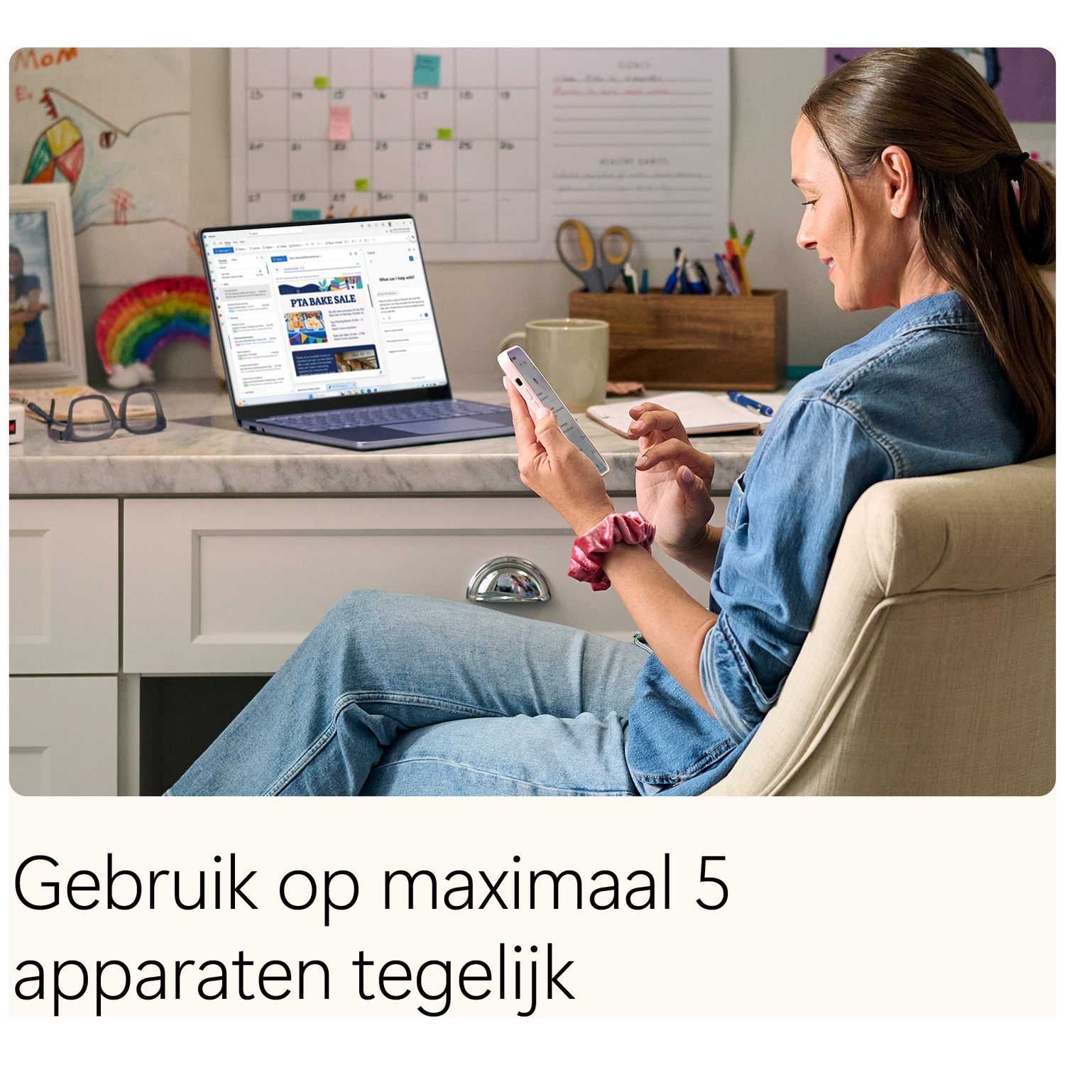 A woman sits at a desk, using her smartphone and looking at an open laptop.