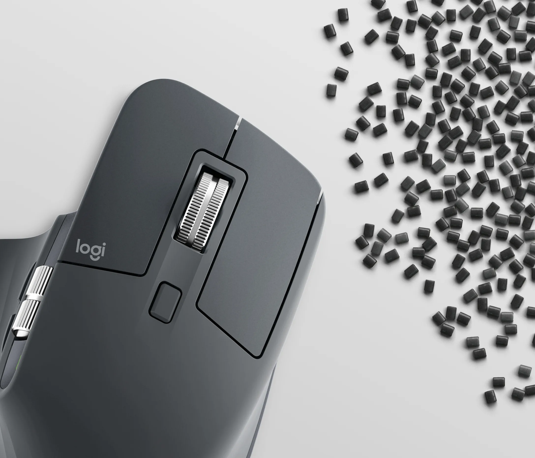 Un mouse wireless Logitech grigio scuro accanto a pellet di plastica nera su superficie bianca.
