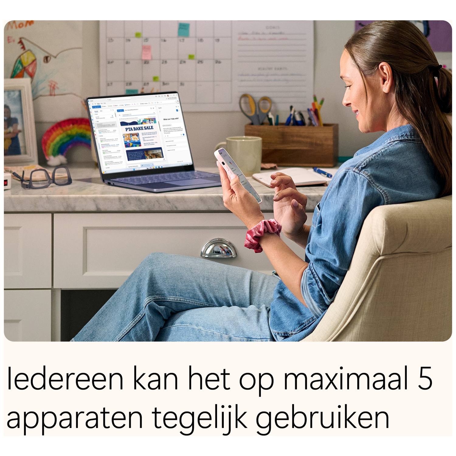 A woman sits at a desk using a smartphone with an open laptop nearby.