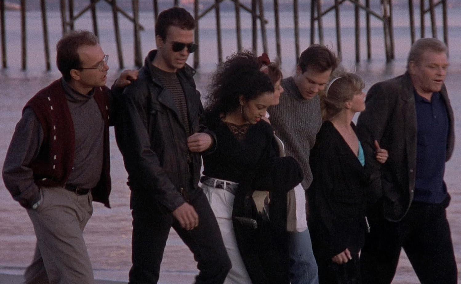 Eine vielfältige Gruppe von Menschen geht am Strand mit hölzernen Piersäulen im Hintergrund.