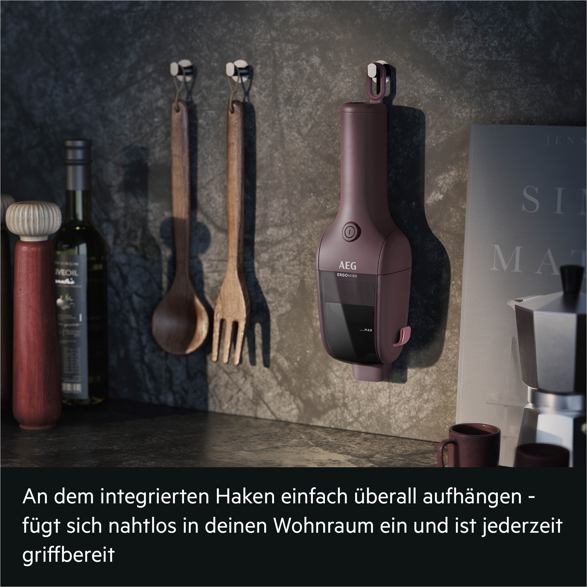 Ein maronifarbener AEG Handstaubsauger hängt an einer dunklen Küchenwand neben Holzlöffeln und einem Buch.