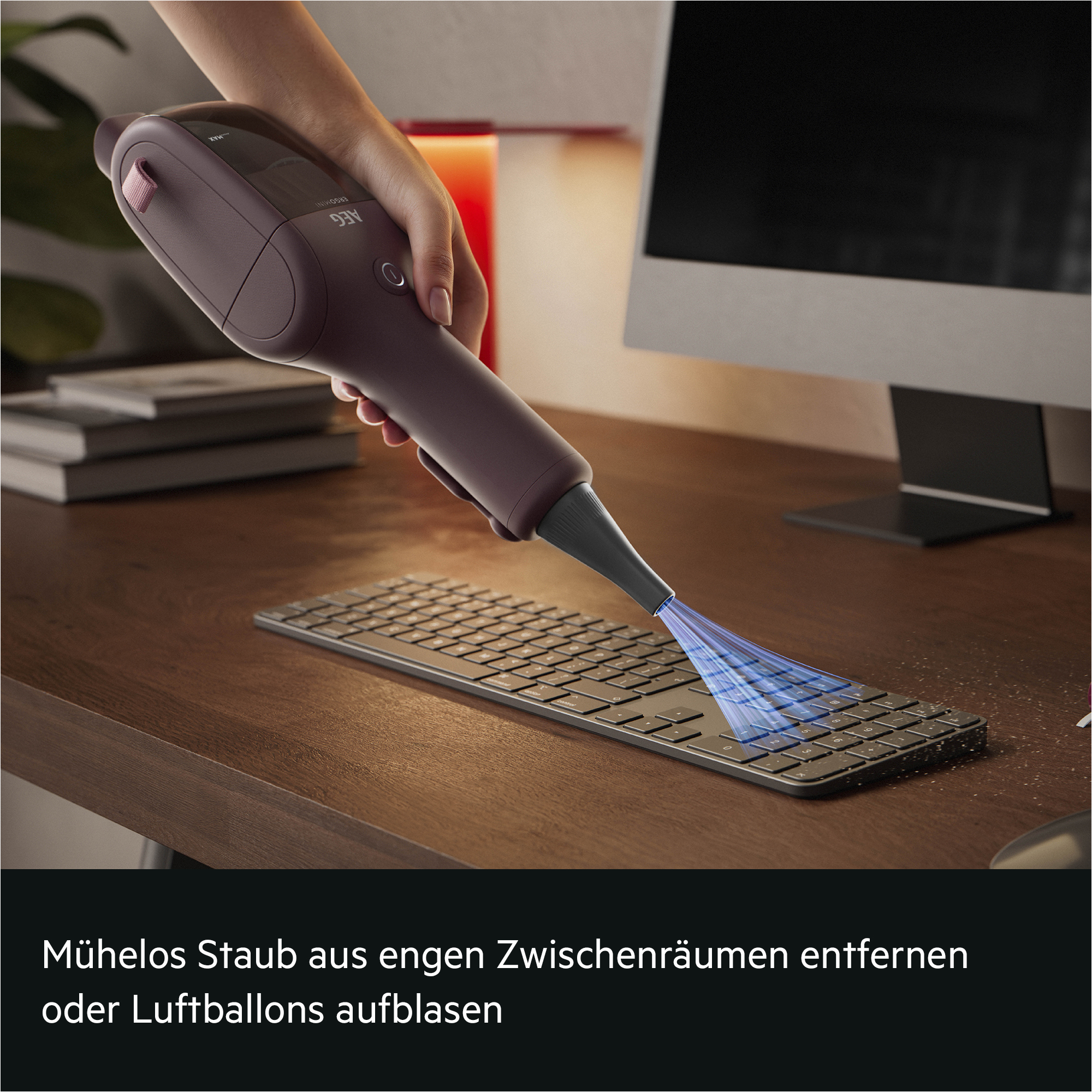 Hand hält einen violetten Akkusauger, der Staub aus einer Computertastatur bläst.