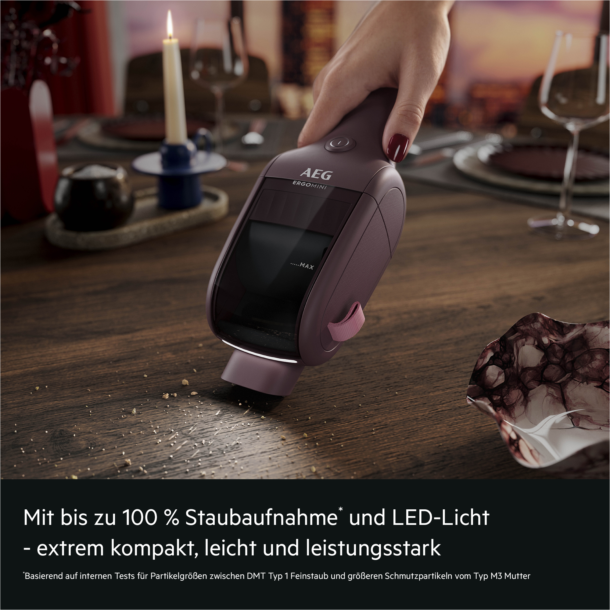 Hand hält einen kompakten, violetten AEG Handstaubsauger, der Krümel von einem Holztisch aufsaugt.