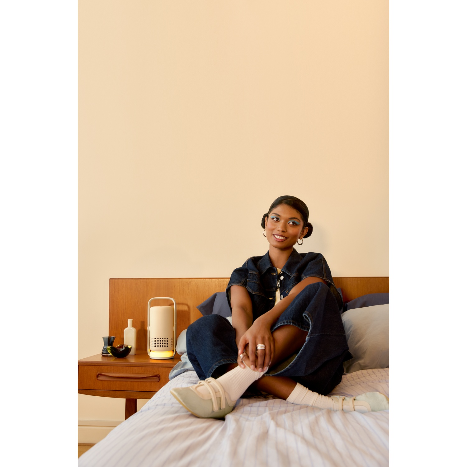 A smiling woman in denim clothes sits on a bed next to a small light.