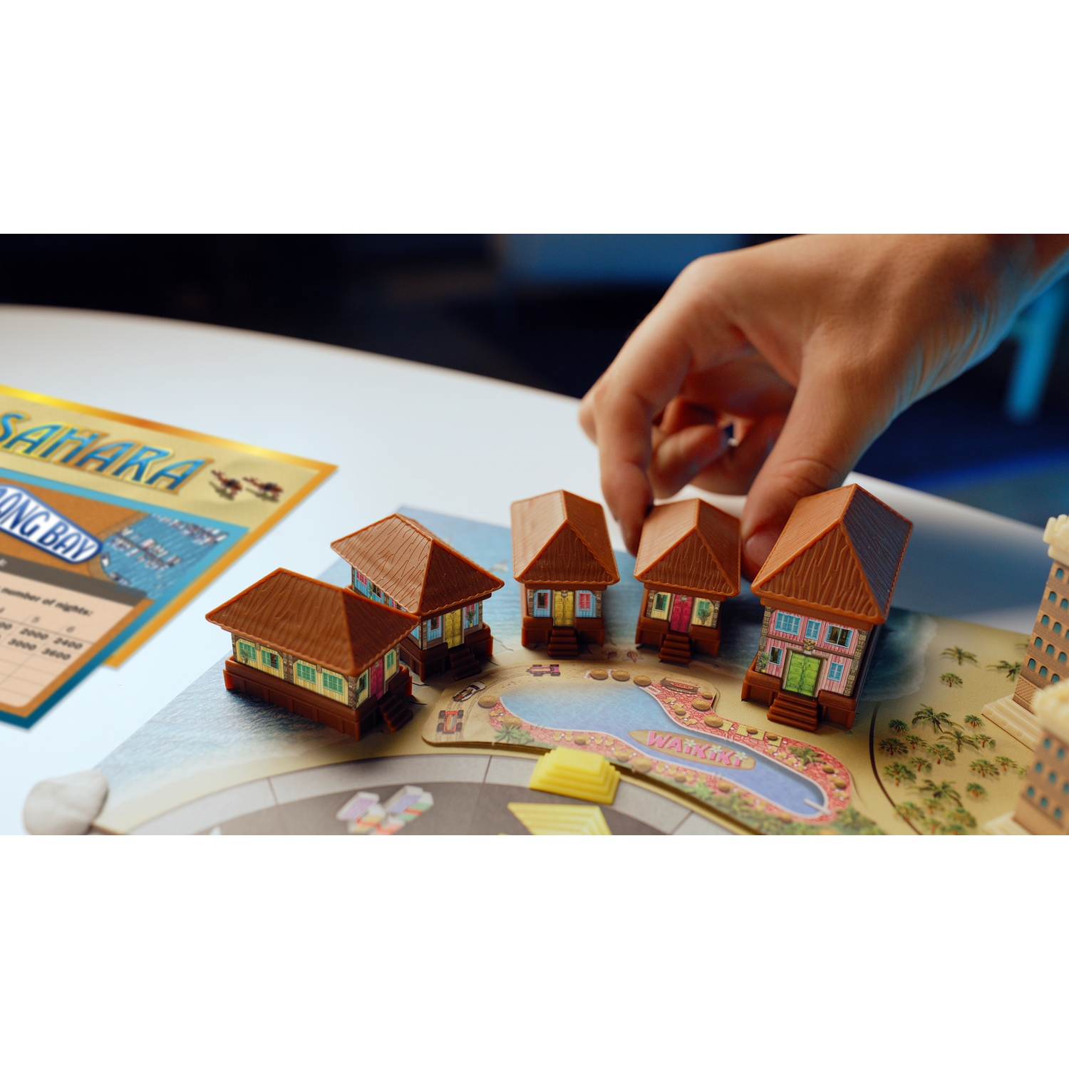 A hand placing small model huts on a board game with a pool and resort theme.