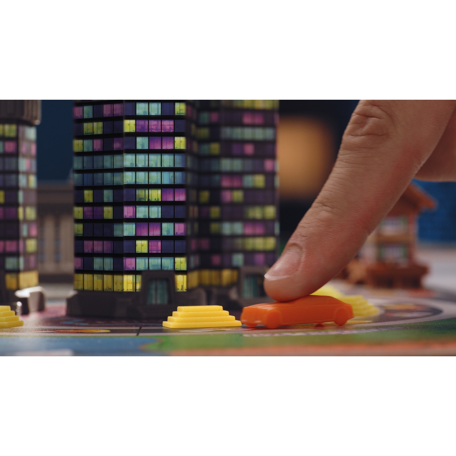A finger moves an orange toy car on a board game with model skyscrapers in the background.
