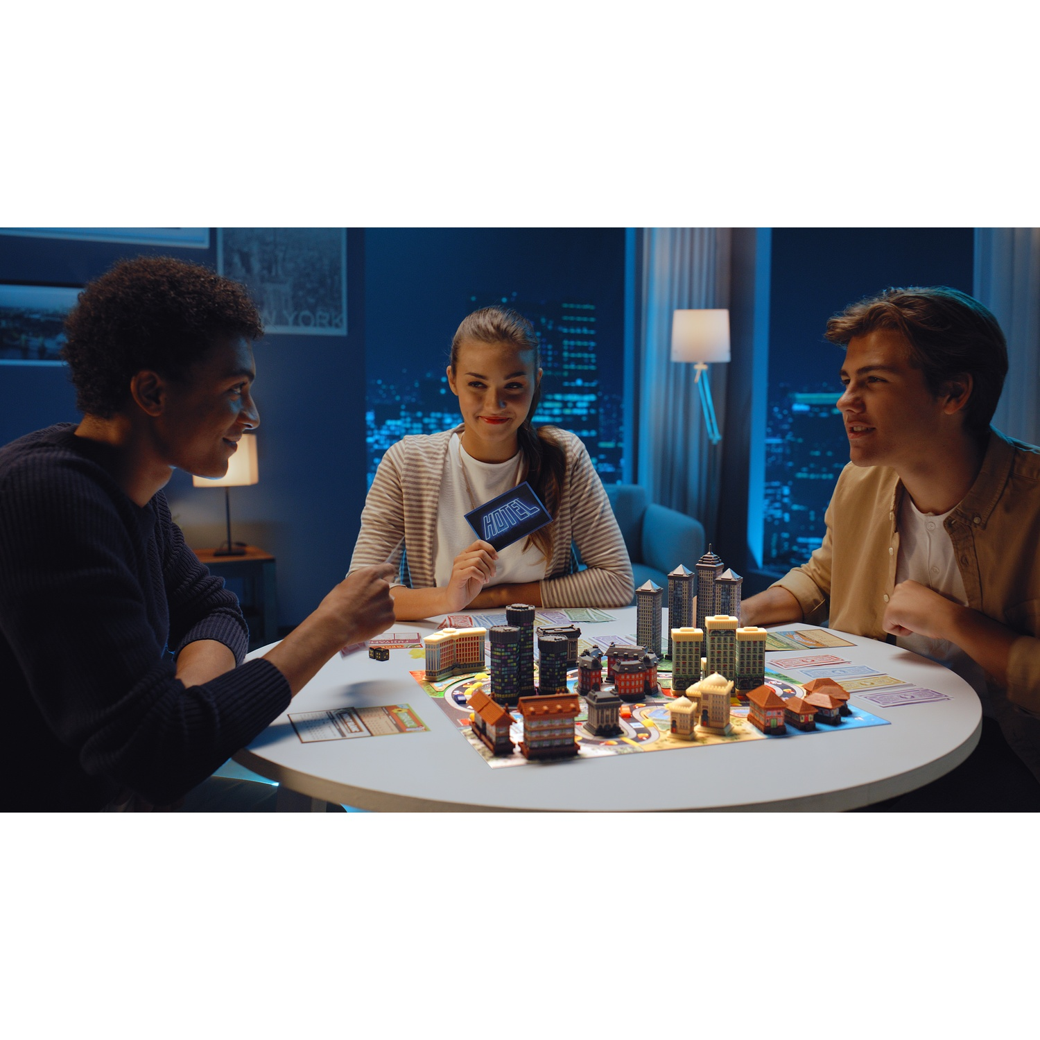 Three young friends play a hotel board game with small buildings at a round table at night.