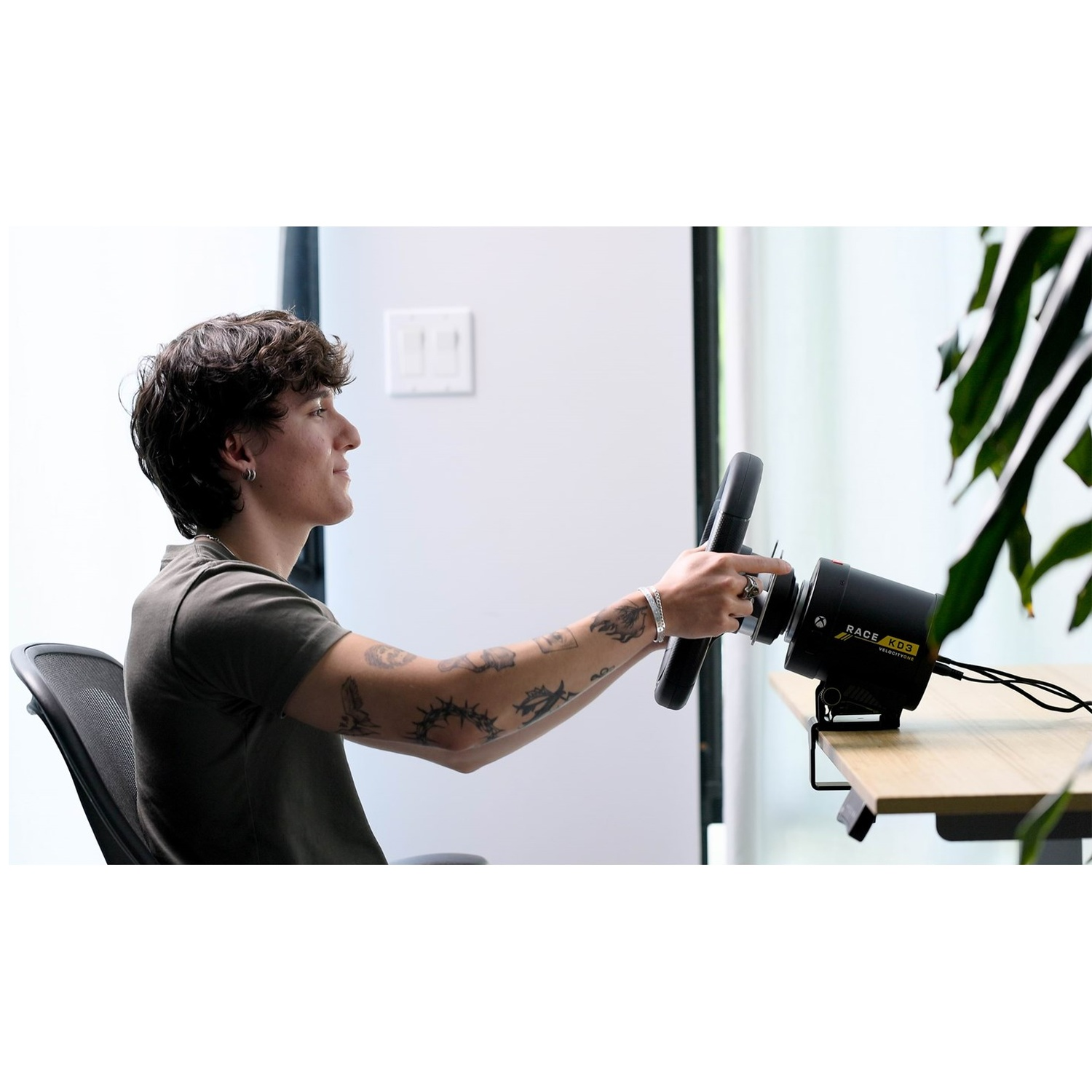 A young man with tattoos sits at a desk, holding and using a racing steering wheel simulator.