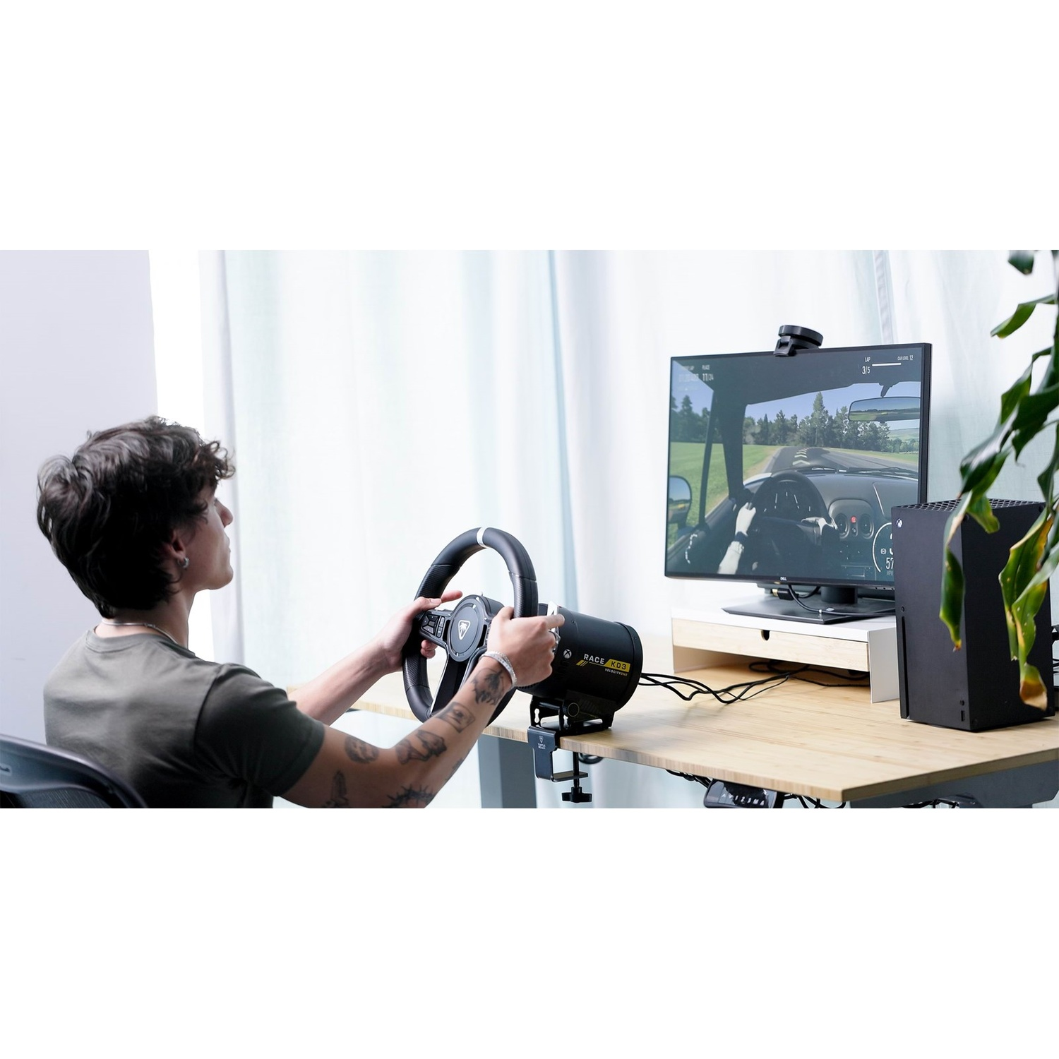 A young man playing a racing game on a monitor, holding a black steering wheel attached to a wooden desk.