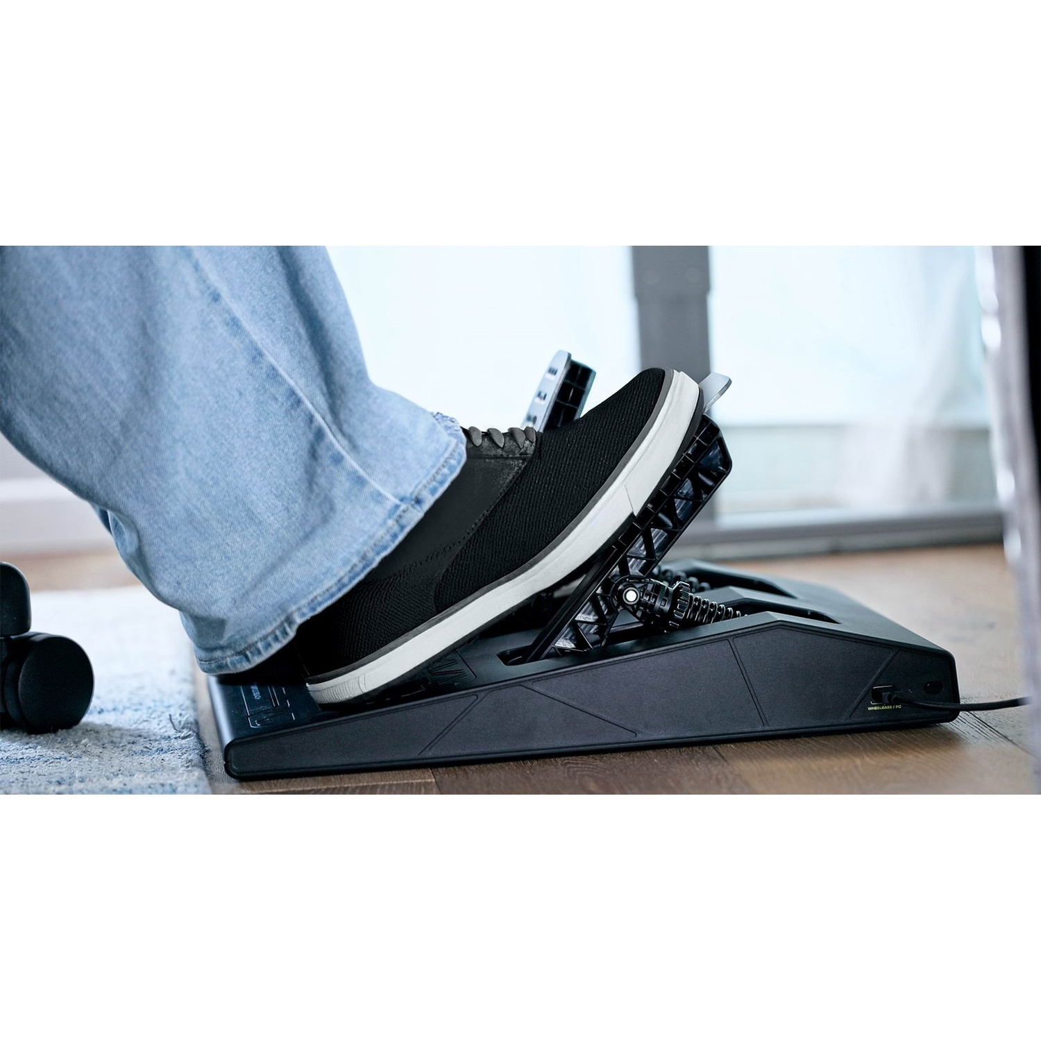 A foot in jeans and black shoe presses on a black metal racing simulator pedal on a wooden floor.