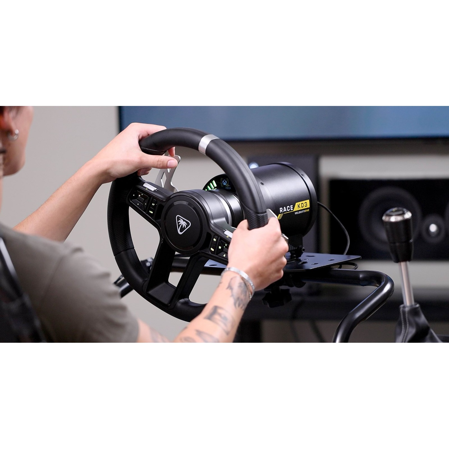 A person holds a black racing steering wheel with buttons and paddles, with a monitor visible in the background.