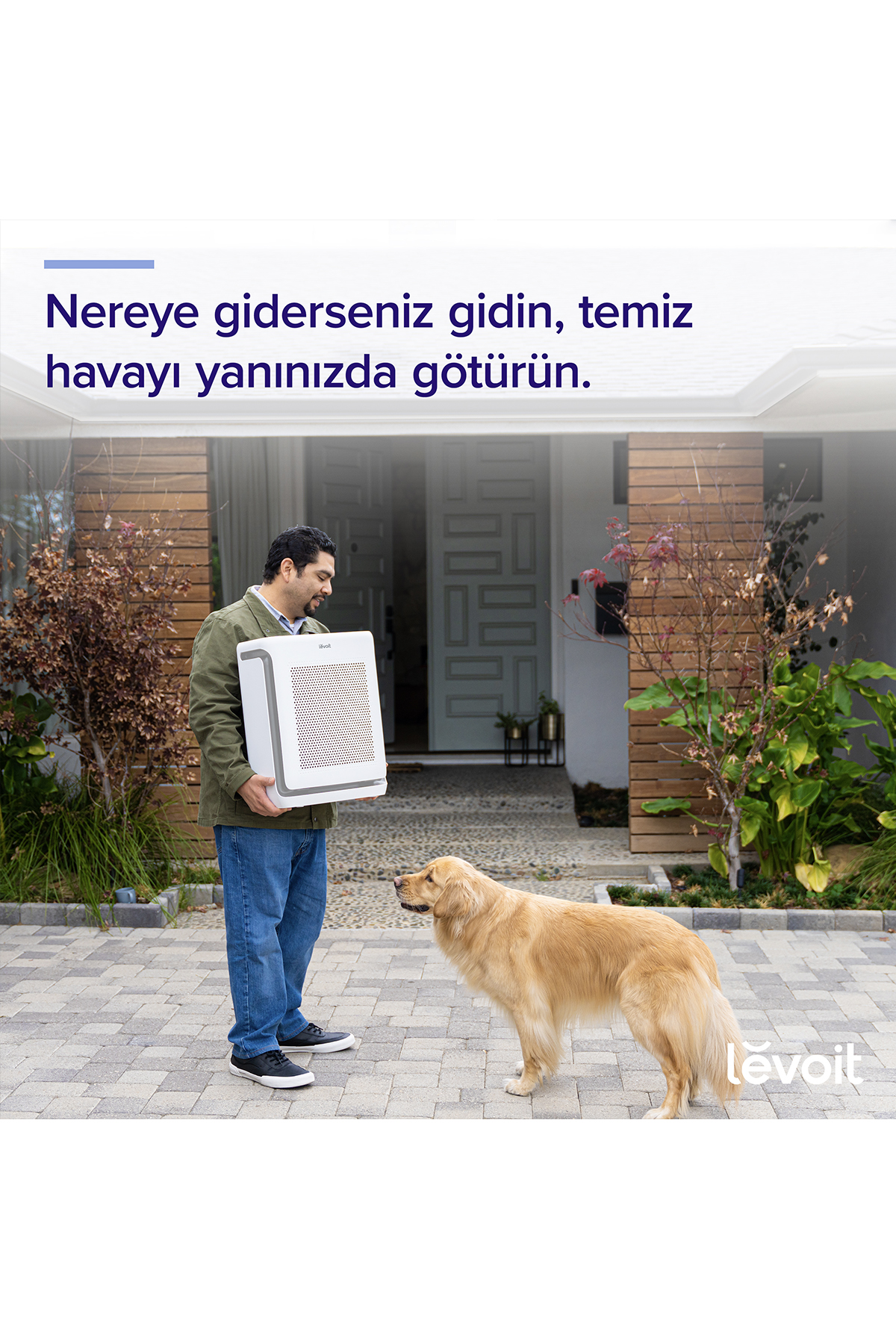 Bir adam evinin önündeki avluda beyaz bir hava temizleyici taşıyor, bir golden retriever ona bakıyor.