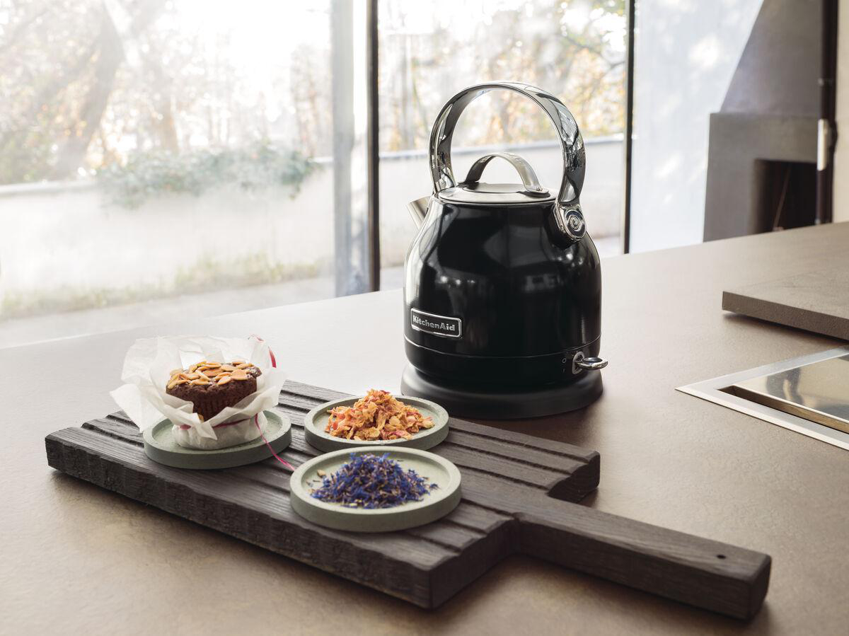 Ein schwarzer KitchenAid Wasserkocher und ein Holzschneidebrett mit Muffin und Teeblättern auf einer Küchenarbeitsplatte.