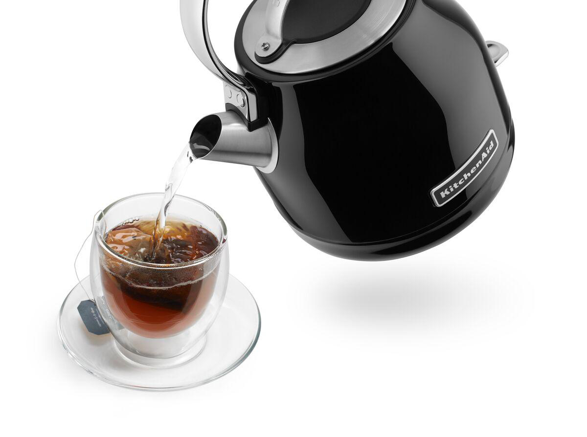 Ein schwarzer KitchenAid-Wasserkocher gießt heißes Wasser in eine Teetasse auf einer Untertasse vor weißem Hintergrund.