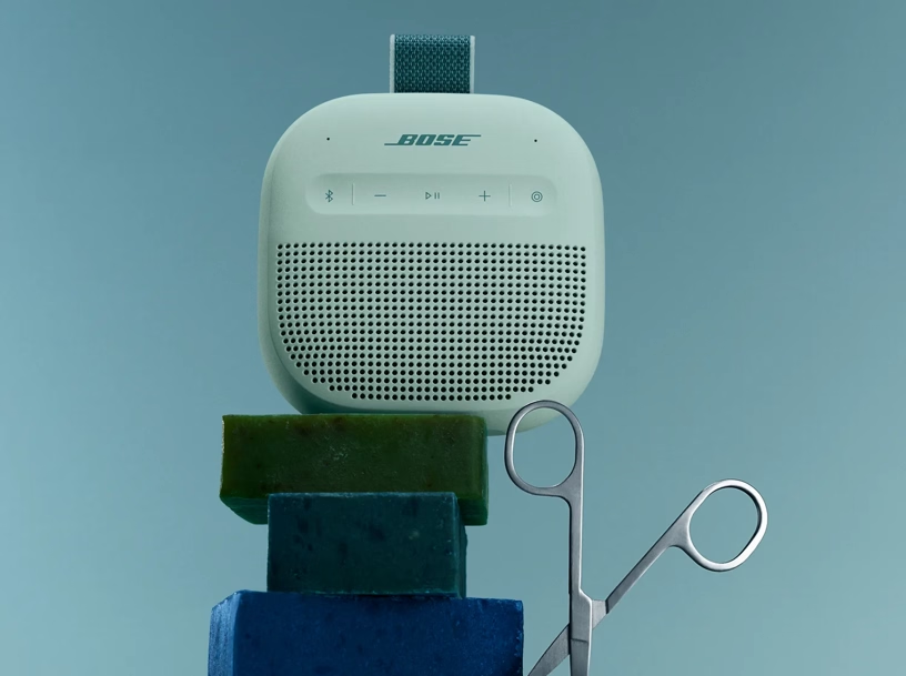 Ein hellgrüner Bose-Lautsprecher auf einer gestapelten Seife, daneben eine Schere auf blauem Hintergrund.
