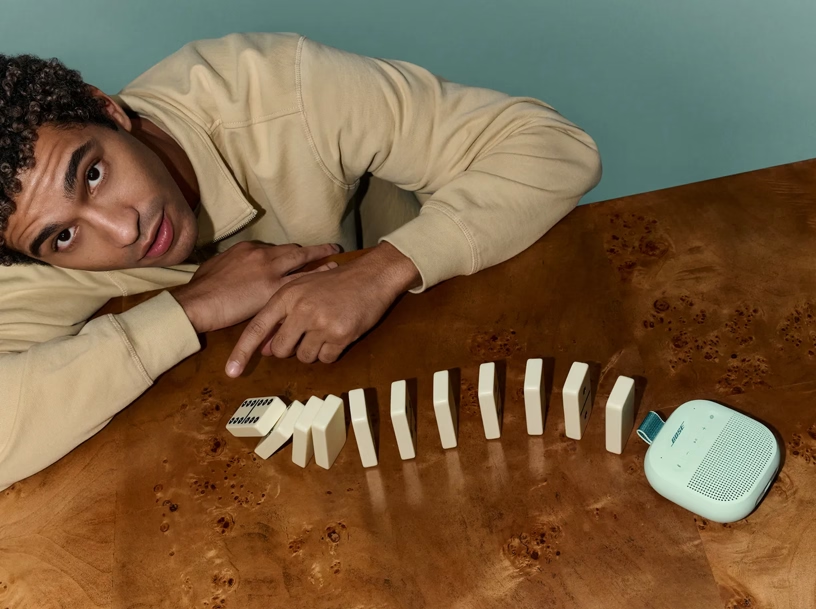 Ein Mann in einem beigen Pullover betrachtet umfallende Dominosteine auf einem Holztisch neben einem hellblauen Lautsprecher.