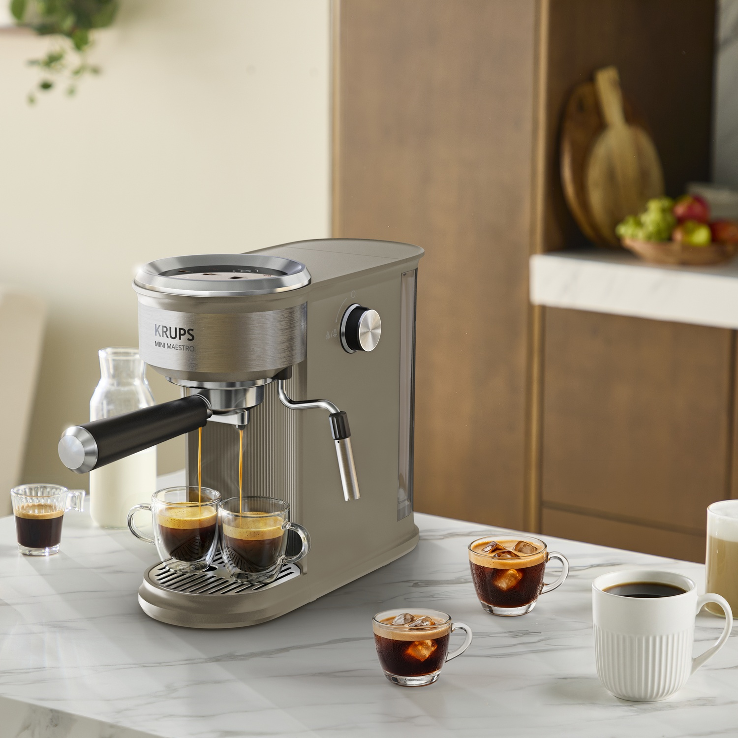 Een grijze Krups Mini Maestro espressomachine schenkt koffie in twee glazen op een marmeren aanrecht.