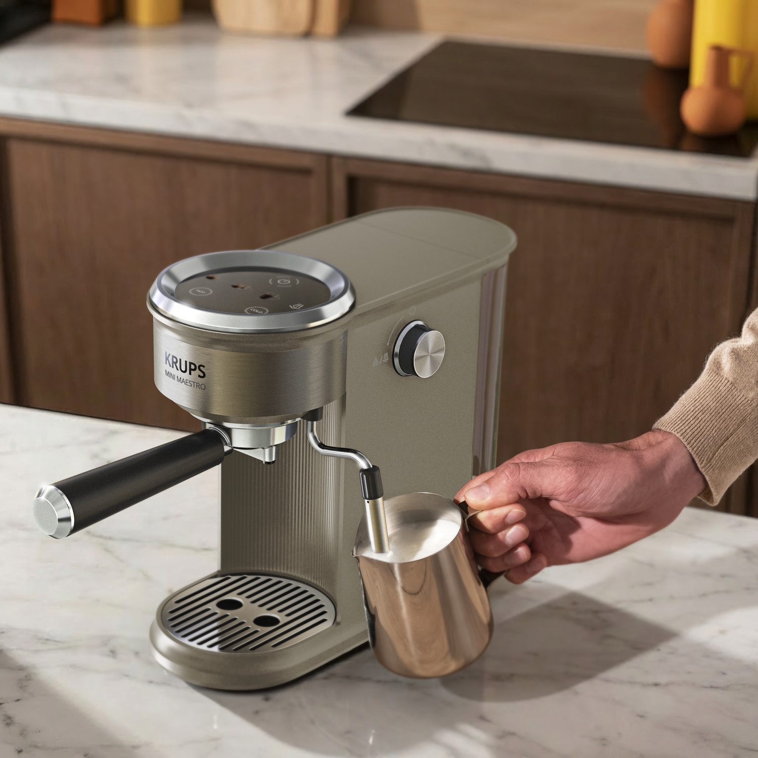 Een hand houdt een melkkan bij het stoompijpje van een gouden Krups espressomachine op een marmeren aanrecht.