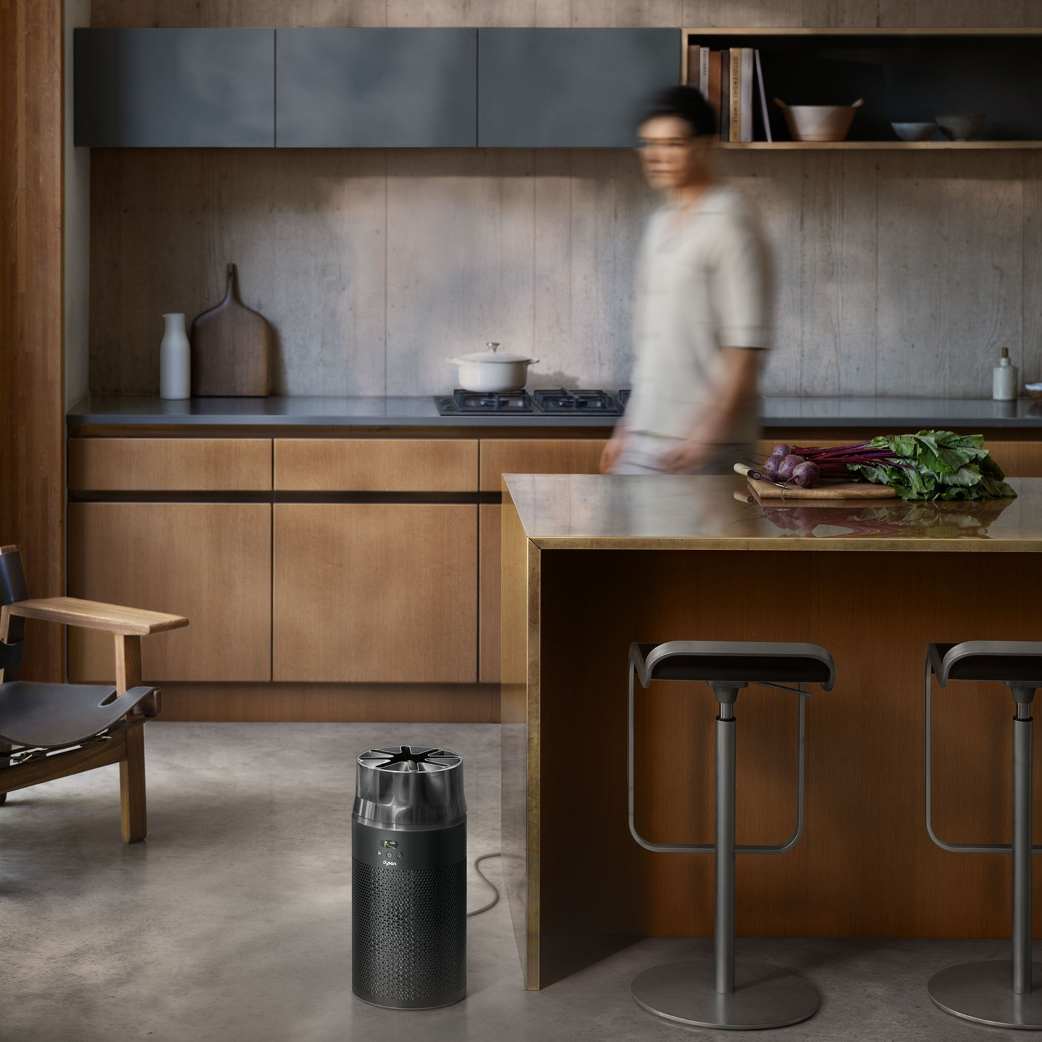 Une cuisine moderne présente un purificateur d'air Dyson noir au premier plan, un îlot en bois et une personne floue.