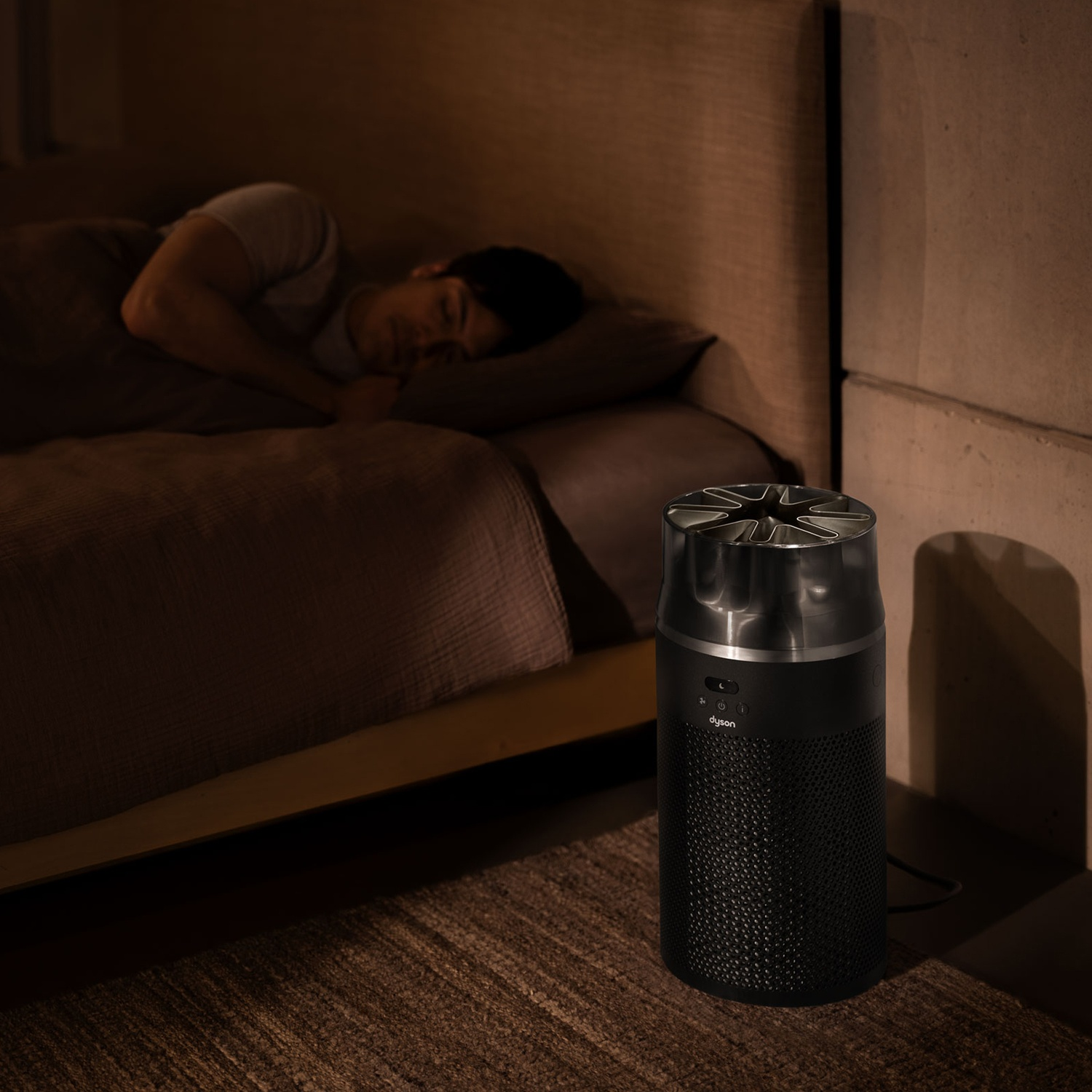 Un purificateur d'air Dyson noir au sol dans une chambre sombre où quelqu'un dort.