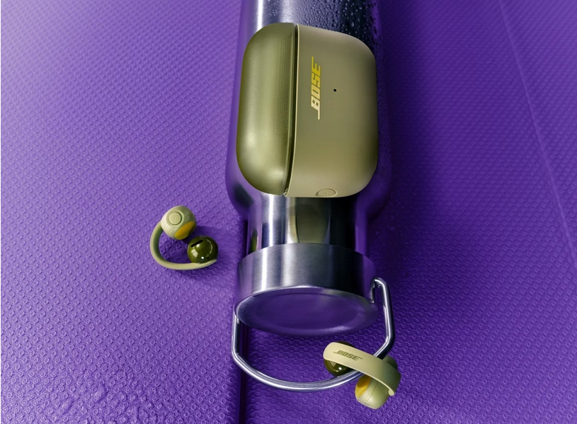 Olivgrüne Bose-Ohrhörer und ein Gehäuse auf einer silbernen Flasche auf einer violetten Matte.