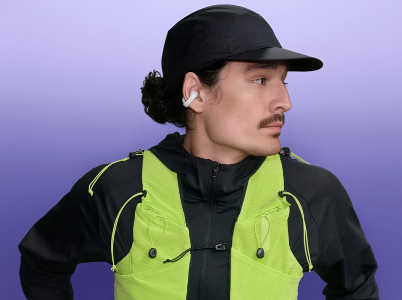 Ein Mann mit schwarzer Mütze, dunkler Jacke und neongrüner Laufweste vor einem einfarbigen lila Hintergrund.