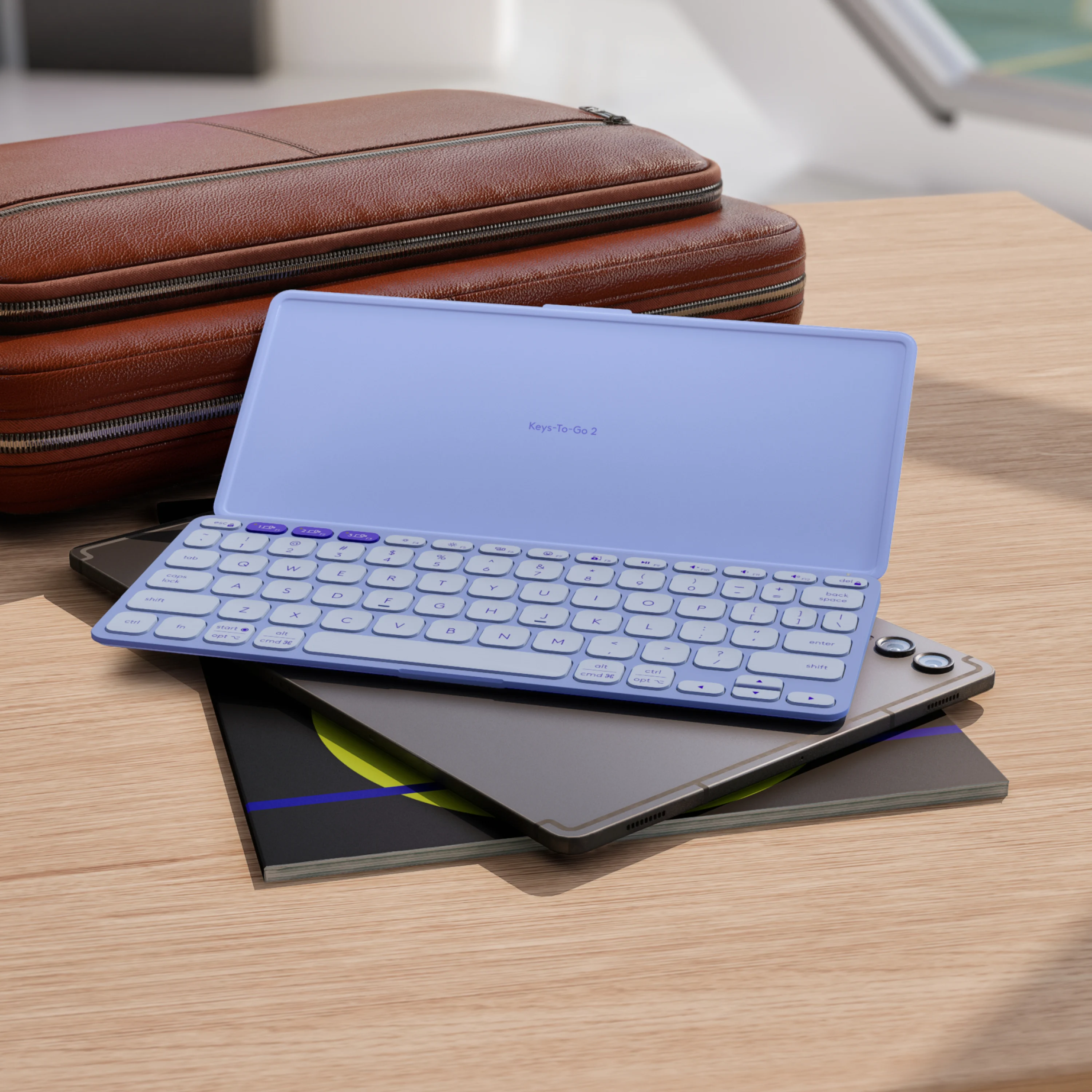 Una tastiera wireless azzurra poggia su tablet e quaderno scuri, con unacustodia in pelle marrone sullo sfondo.