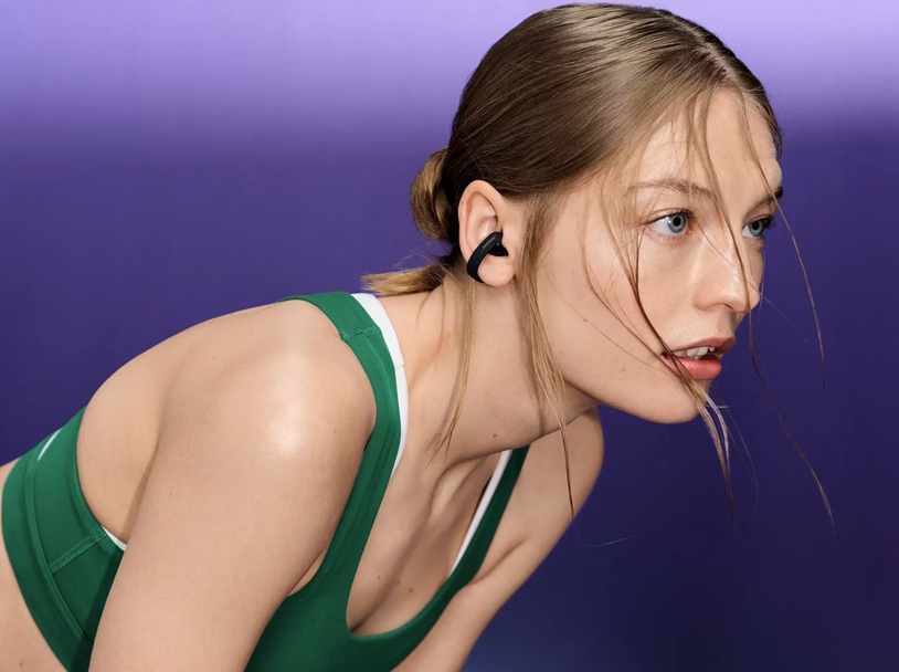 Frau mit offenem Haar und grünem Sport-Top trägt einen schwarzen In-Ear-Kopfhörer vor violettem Hintergrund.