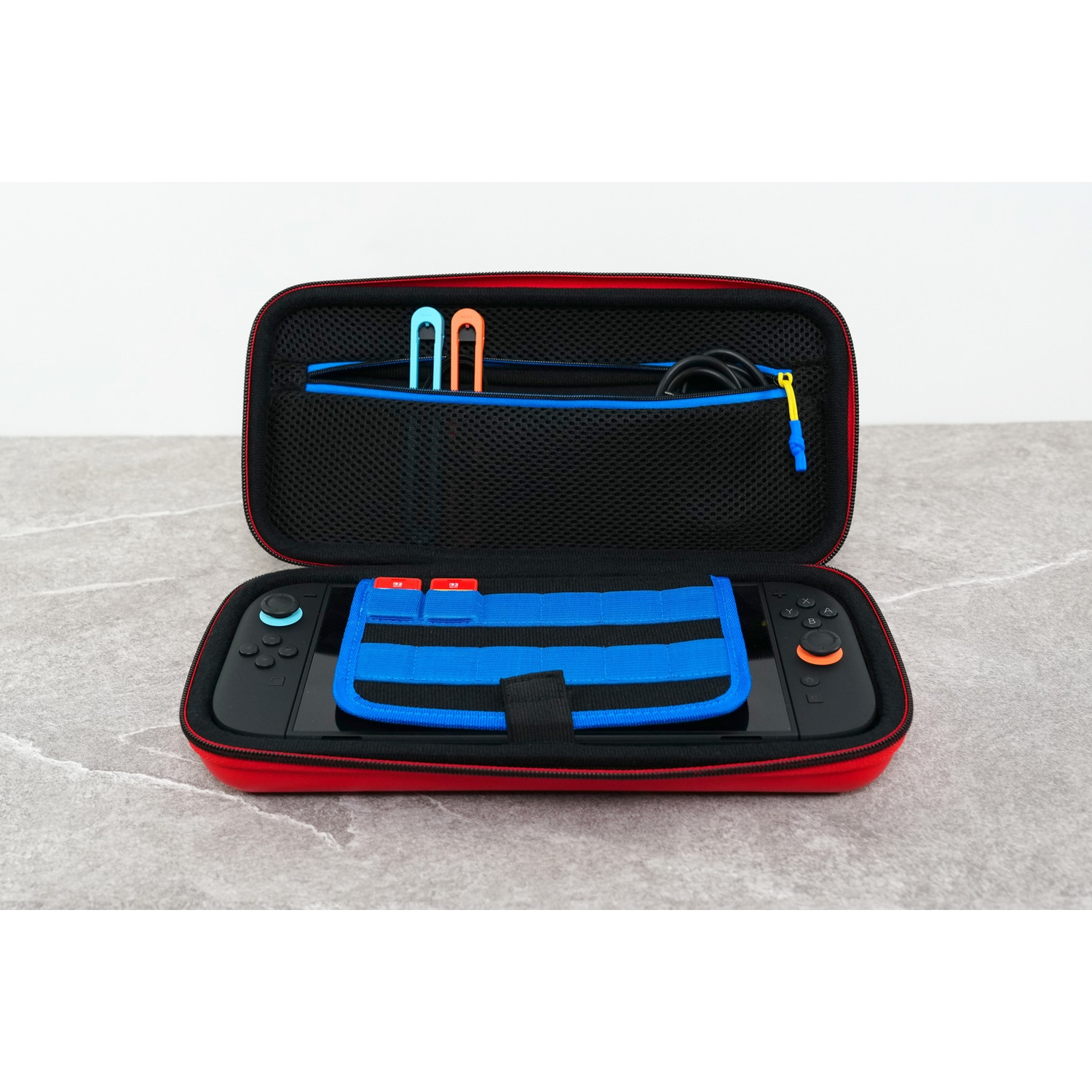 An open red and black gaming console case on a grey surface, holding a black console and accessories.