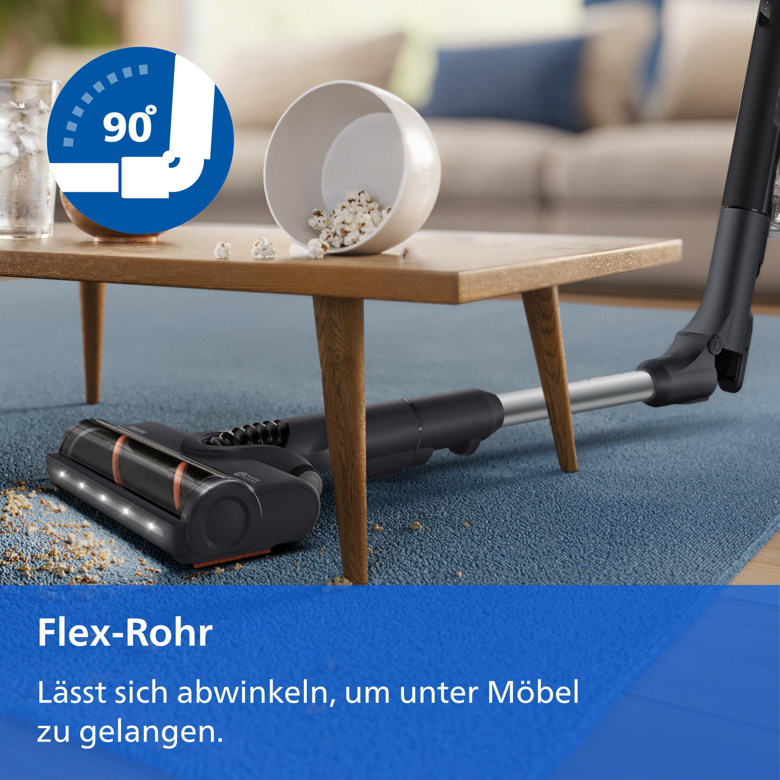 Ein Staubsauger mit Flex-Rohr reinigt unter einem Couchtisch auf blauem Teppich mit verschüttetem Popcorn.