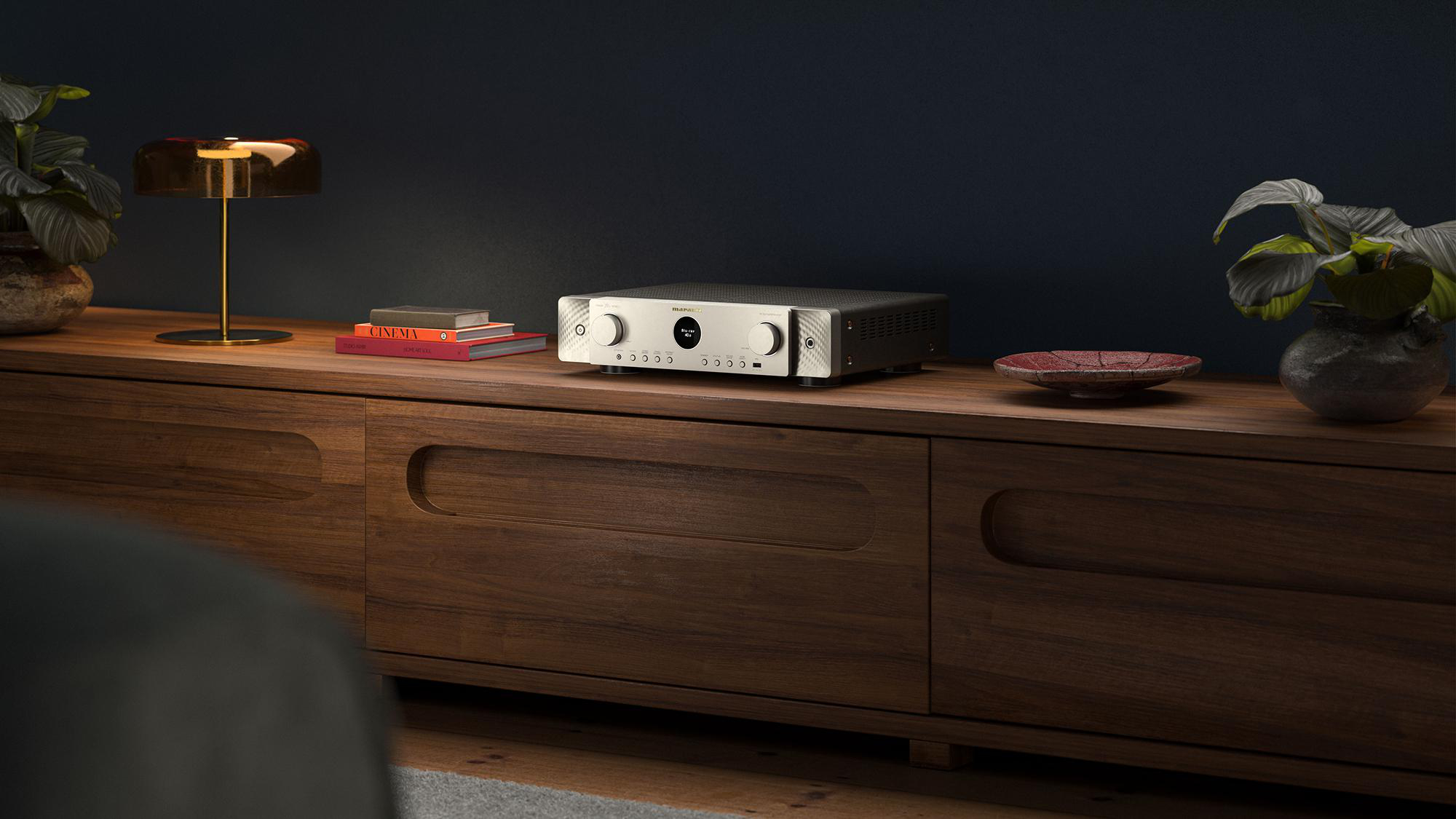 Ein silberner AV-Receiver auf einem Holzbüfett, flankiert von Lampen, Büchern und Topfpflanzen vor blauer Wand.