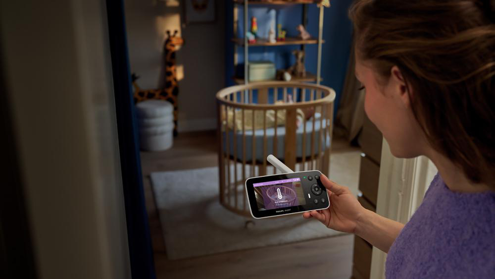 Eine Frau hält ein Babyphone, das den Raum mit einem runden Kinderbett zeigt.