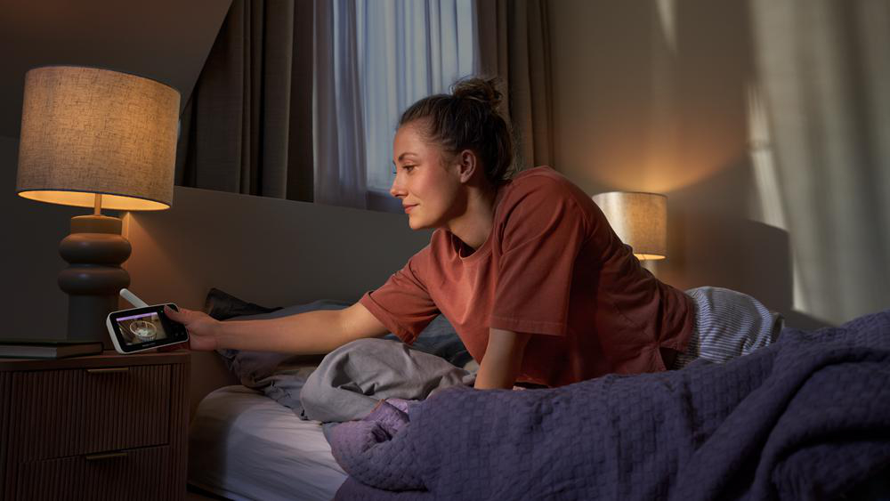 Eine Frau in einem Schlafzimmer hält ein Babyphone in der Hand, während sie auf dem Bett kniet.
