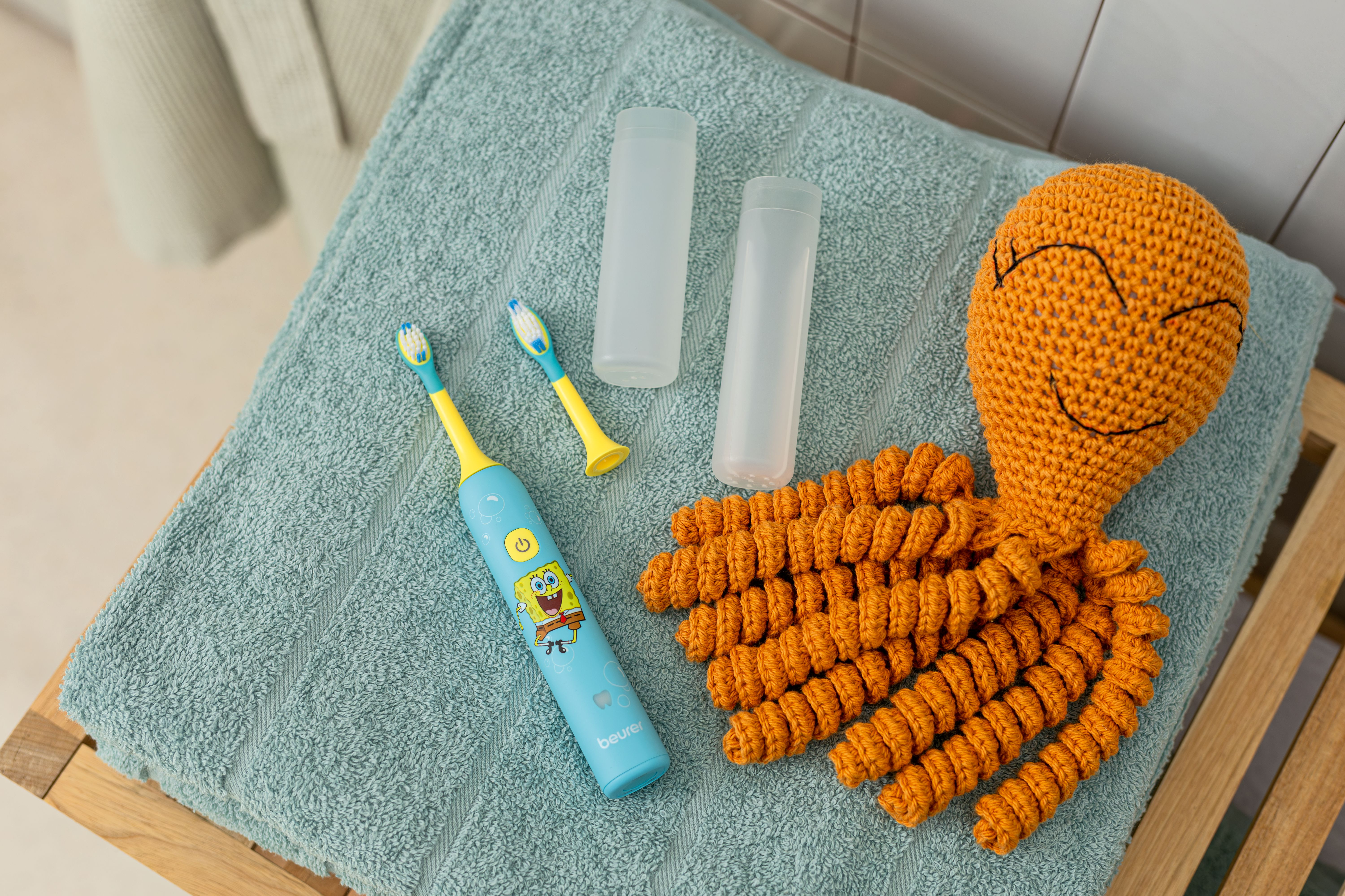 Eine elektrische Kinderzahnbürste mit SpongeBob, zwei Etuis und ein gehäkelter Oktopus liegen auf einem grünen Handtuch.