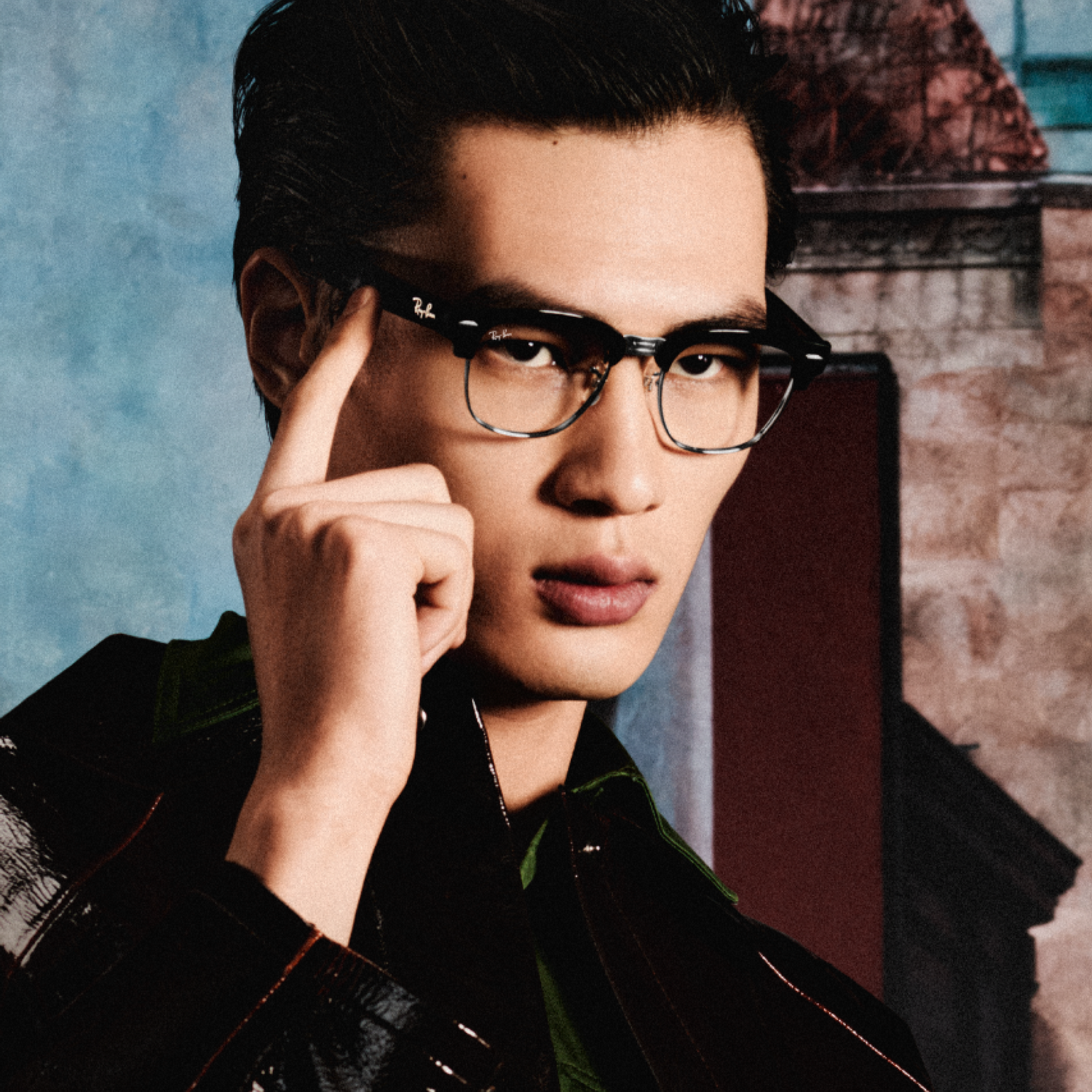 Un joven con gafas toca la montura con la mano frente a un fondo pintado.
