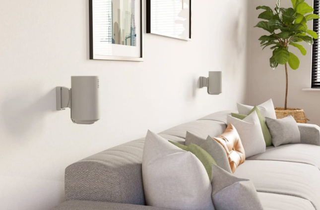 Zwei graue Lautsprecher an Wand montiert über einem Sofa mit Kissen in hellem Wohnzimmer.