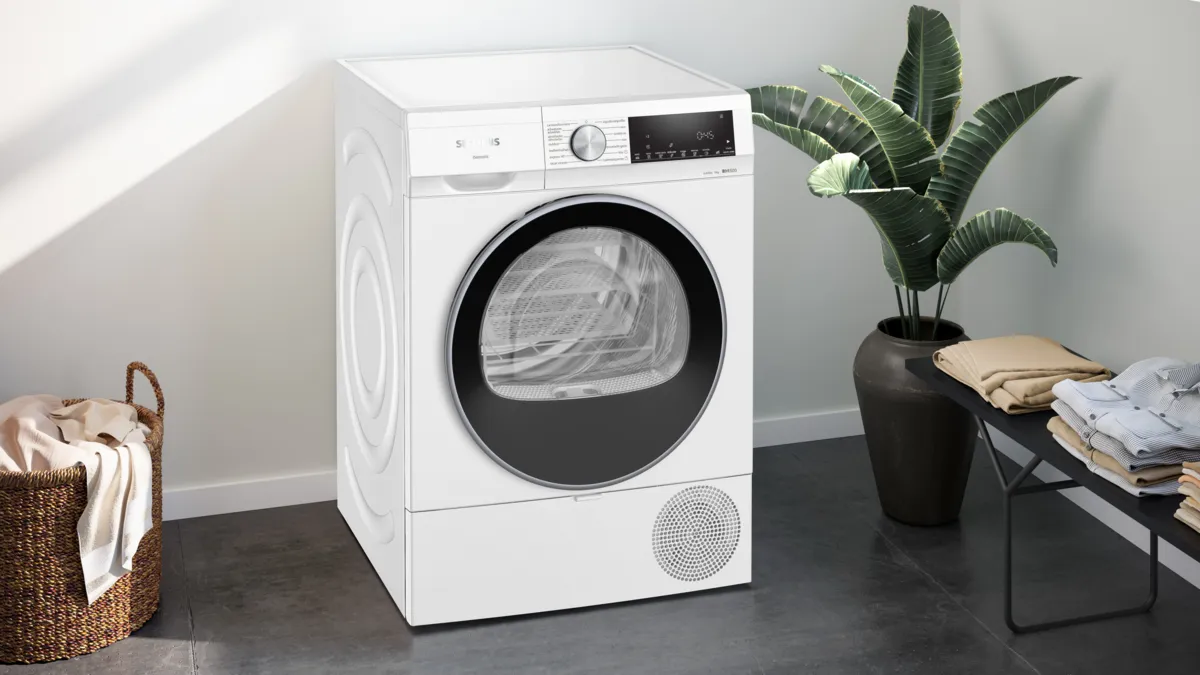 Una secadora blanca en una habitación luminosa, junto a un cesto, una planta y ropa doblada.